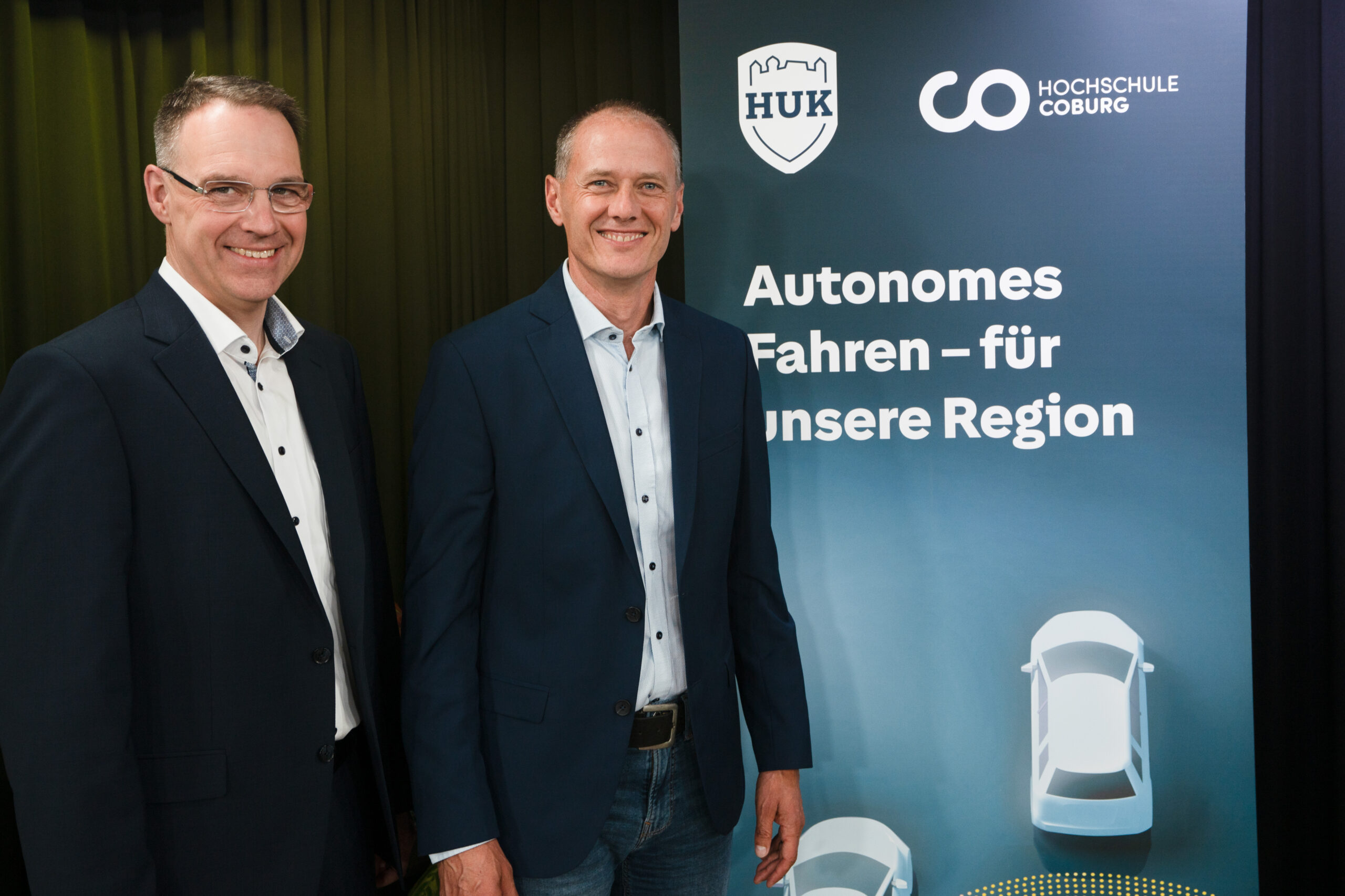 Zwei lächelnde Männer in Anzügen stehen neben einem Schild mit Logos und einem deutschen Text zum Thema „autonomes Fahren“ in der Region. Das Schild, das mit den Logos von HUK-COBURG und der Hochschule Coburg versehen ist, zeigt Autosymbole, die für Fortschritte in der Technologie des autonomen Fahrens stehen. at Hochschule Coburg