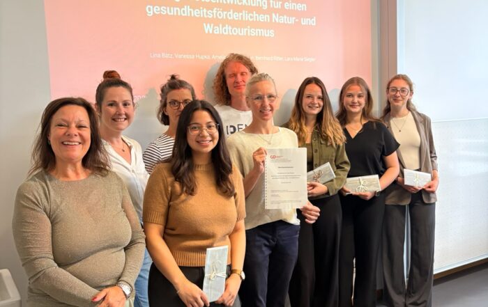 Neun Personen stehen in einem Raum und lächeln in die Kamera; einige halten Zertifikate in den Händen. Hinter ihnen ist eine Präsentationsfolie in deutscher Sprache zu sehen, die sich mit Naturtourismus und gesundheitsförderndem Tourismus an der Hochschule Coburg befasst, wobei insbesondere auf Projekte in Bad Rodach und im Sinneswald eingegangen wird. at Hochschule Coburg