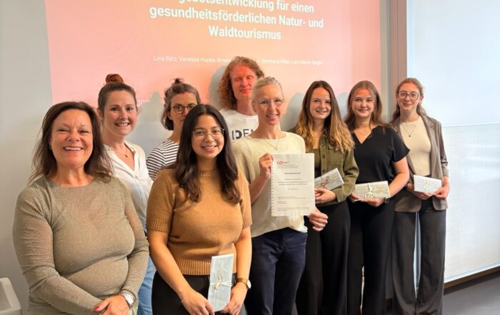 Neun Personen stehen in einem Raum und lächeln in die Kamera; einige halten Zertifikate in den Händen. Hinter ihnen ist eine Präsentationsfolie in deutscher Sprache zu sehen, die sich mit Naturtourismus und gesundheitsförderndem Tourismus an der Hochschule Coburg befasst, wobei insbesondere auf Projekte in Bad Rodach und im Sinneswald eingegangen wird. at Hochschule Coburg