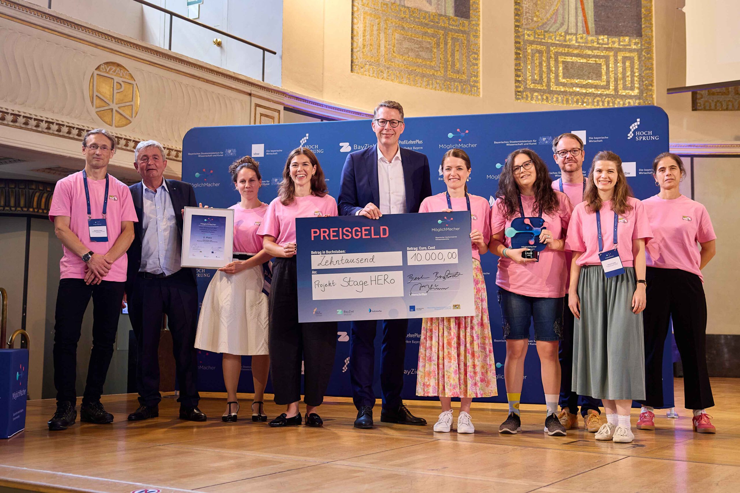 Bei einer Preisverleihung posiert eine Gruppe von Menschen auf der Bühne mit einem großen Scheck über 10.000 €, einer Plakette und einer Trophäe. Die meisten tragen einheitliche rosa Hemden. Im Hintergrund sind Veranstaltungsbanner und architektonische Details zu sehen, während zwischen den Ausstellungsstücken ein „Automatisch gespeicherter Entwurf“ zu erkennen ist. at Hochschule Coburg