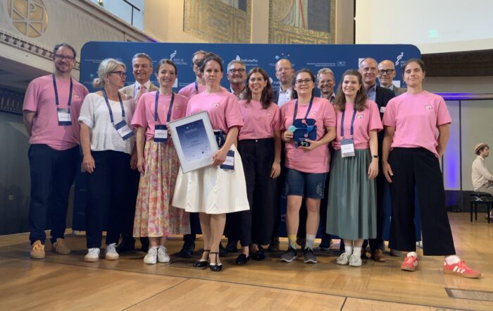 Eine Gruppe von Menschen, von denen einige rosa Hemden tragen, steht auf einer Bühne und lächelt für ein Foto. Einer hält eine gerahmte Urkunde mit der Aufschrift „Automatisch gespeicherter Entwurf“ in der Hand, während ein anderer eine blaue Trophäe präsentiert. Im Hintergrund sind ein blaues Banner und verzierte Wände zu sehen. at Hochschule Coburg