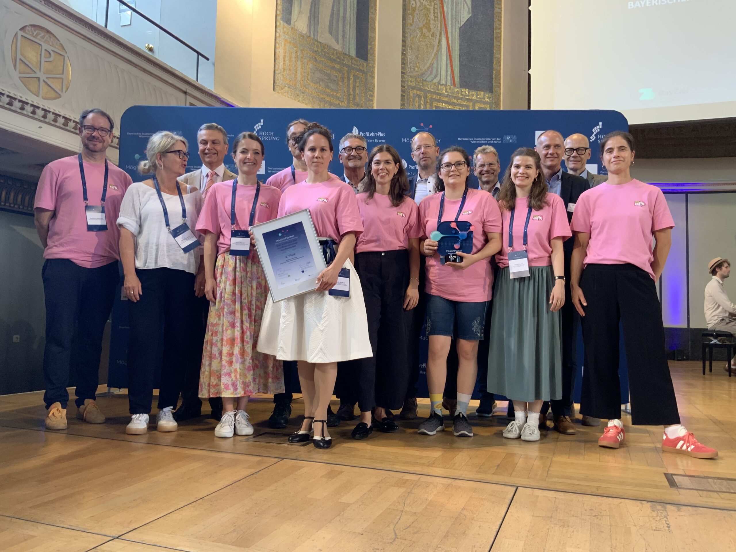 Eine Gruppe von Menschen, von denen einige rosa Hemden tragen, steht auf einer Bühne und lächelt für ein Foto. Einer hält eine gerahmte Urkunde mit der Aufschrift „Automatisch gespeicherter Entwurf“ in der Hand, während ein anderer eine blaue Trophäe präsentiert. Im Hintergrund sind ein blaues Banner und verzierte Wände zu sehen. at Hochschule Coburg