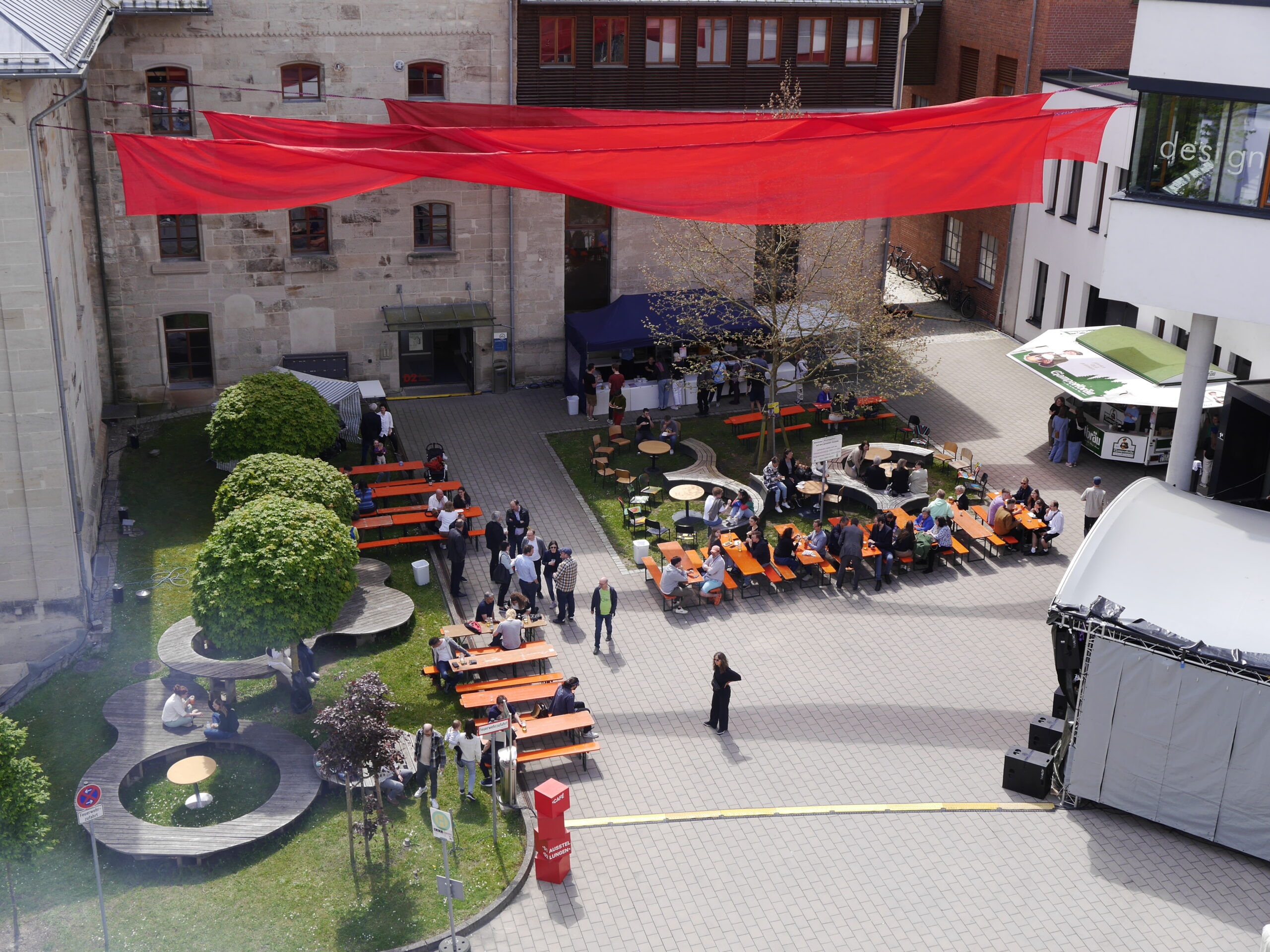 Eine Veranstaltung im Innenhof mit Menschen, die auf orangefarbenen Bänken sitzen, einer Bühne, Imbissständen und über den Köpfen aufgehängten roten Stoffbahnen. Bäume und Grünflächen umgeben den gepflasterten Versammlungsplatz und schaffen eine einladende Atmosphäre für Gespräche über den Karriereweg und Möglichkeiten wie die Professur. at Hochschule Coburg