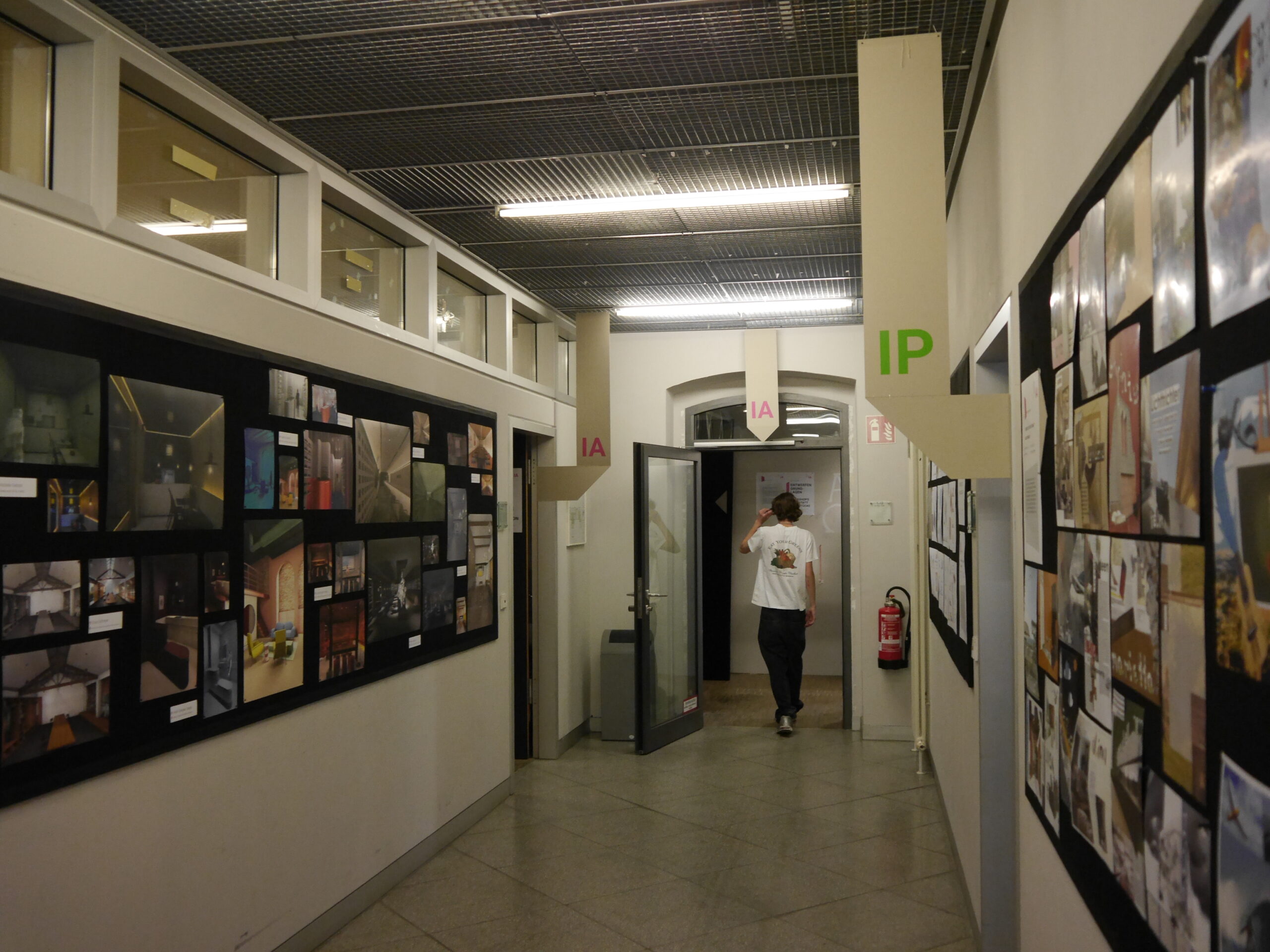 Eine Person geht einen Flur entlang, der auf beiden Seiten von Kunstausstellungen gesäumt ist, die die vielfältigen Wege entlang eines Karrierewegs zur Professur widerspiegeln. Schilder mit den Aufschriften „1A“ und „1P“ hängen von der Decke über dem grauen Boden und der Metallgitterdecke und leiten die Besucher durch diesen kreativen akademischen Korridor. at Hochschule Coburg