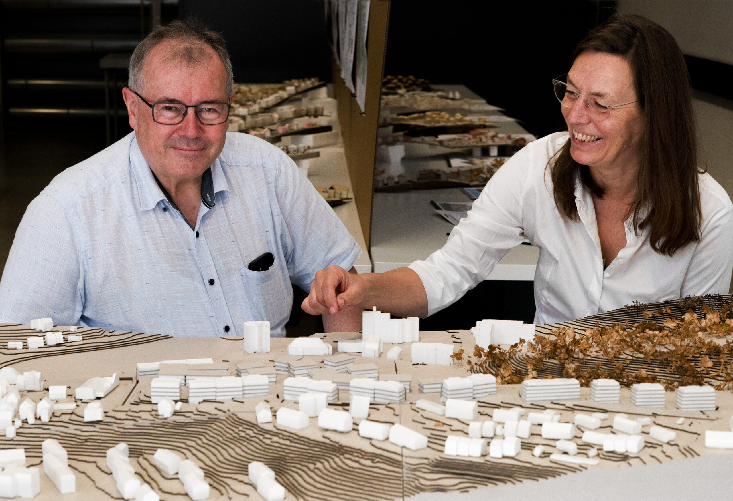 Zwei Menschen lächeln und zeigen auf ein detailreiches Architekturmodell mit weißen Gebäuden und braunen Landschaftselementen, das vor ihnen auf einem Tisch ausgebreitet ist. Dabei fangen sie den kreativen Geist der Werkschau der Fakultät Design ein, während sie über innovative Ansätze im Bauen diskutieren. at Hochschule Coburg