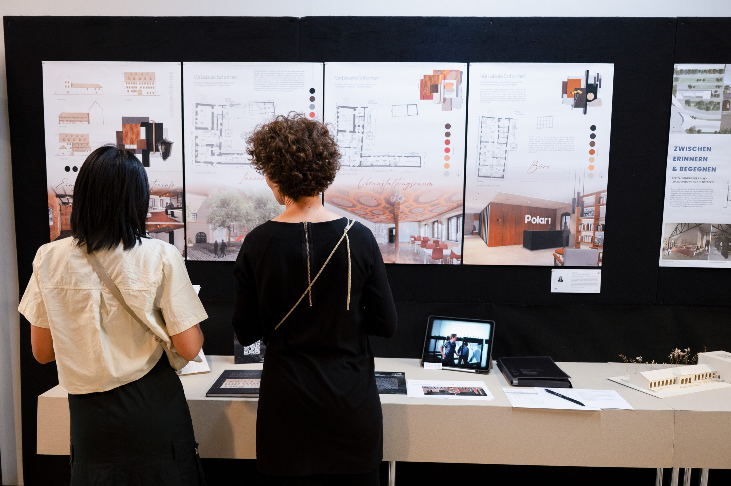 Zwei Personen stehen vor einer Ausstellungstafel auf der Werkschau und betrachten Architekturpläne und Entwürfe der Fakultät Design. Auf dem Tisch vor ihnen liegen außerdem ein Tablet, ein Buch und ein kleines Modell zum Thema „Bauen“. at Hochschule Coburg