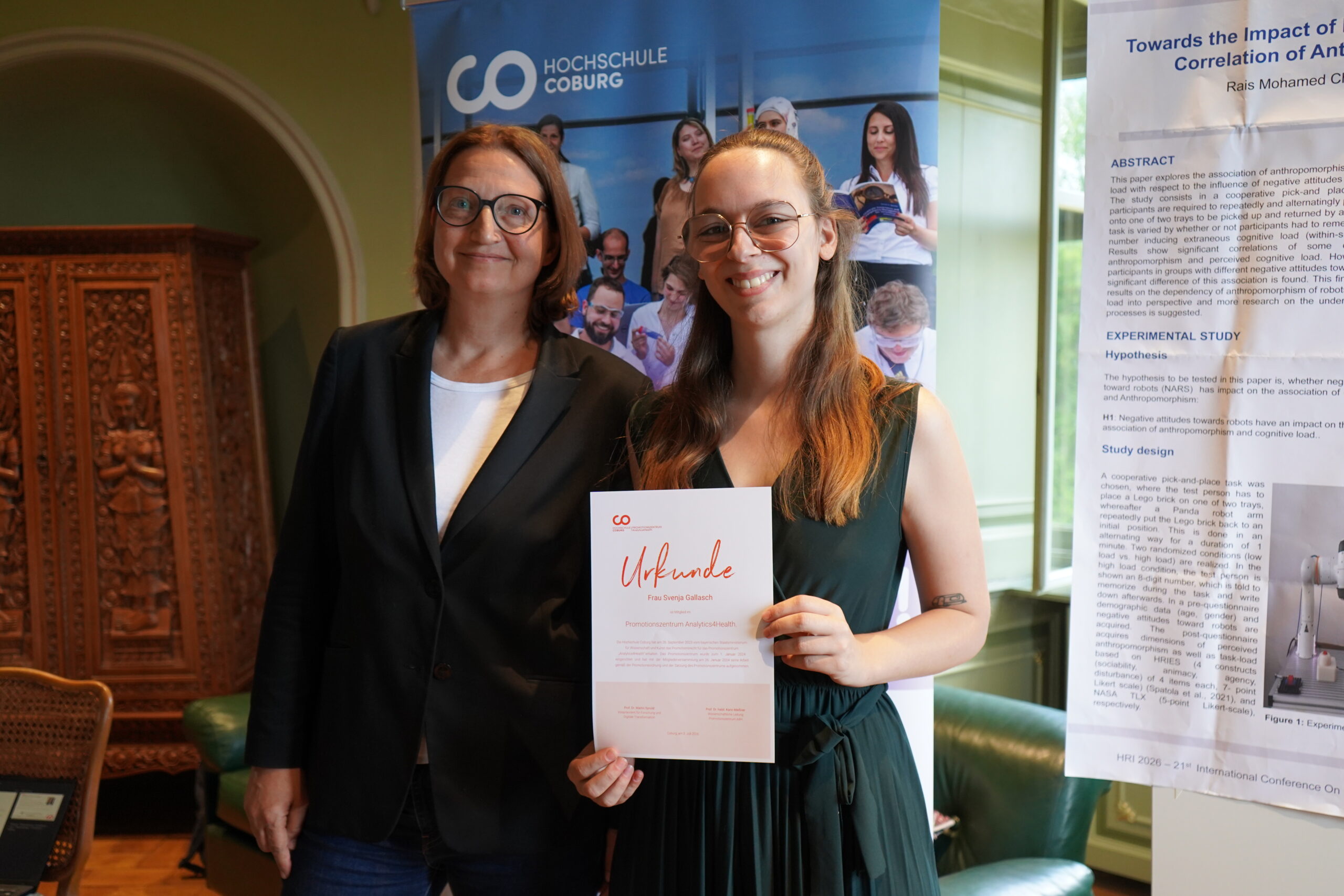 Zwei Frauen stehen in einem Raum, eine von ihnen hält eine Urkunde in der Hand und lächelt. Im Hintergrund sind ein Banner der Hochschule Coburg und ein Forschungsplakat zu sehen, die den Geist des Austauschs bei einer Promotionstagung für gute Doktorarbeiten unterstreichen. at Hochschule Coburg