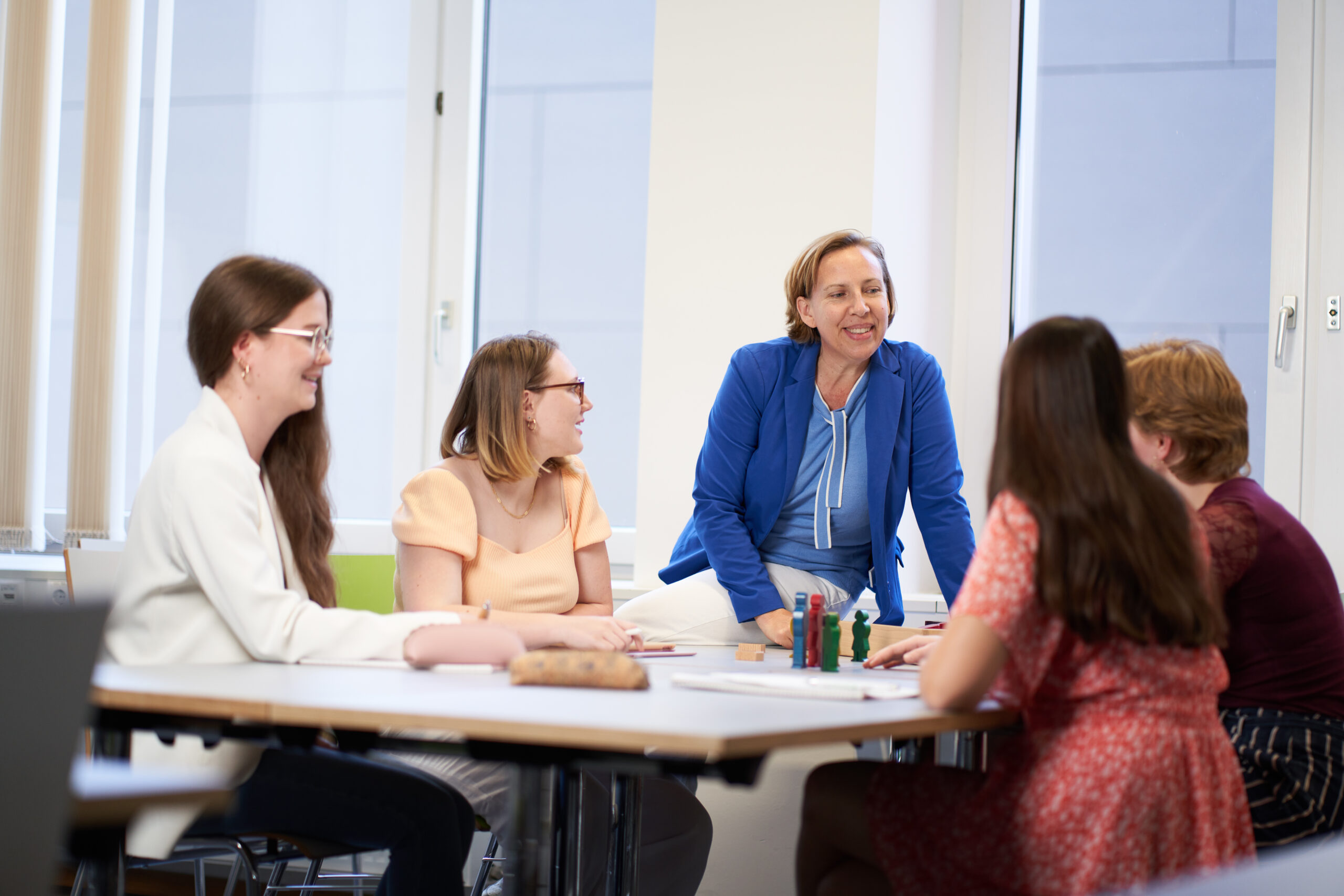 Vier Frauen sitzen lächelnd und plaudernd um einen Tisch herum, während eine weitere Frau in einem blauen Blazer in einem hellen, modernen Büro oder Unterrichtsraum steht und sich mit ihnen unterhält. Die motivierende Atmosphäre lässt auf ein anregendes Gespräch über ihre akademische Laufbahn und den Karriereweg zur Professur schließen. at Hochschule Coburg