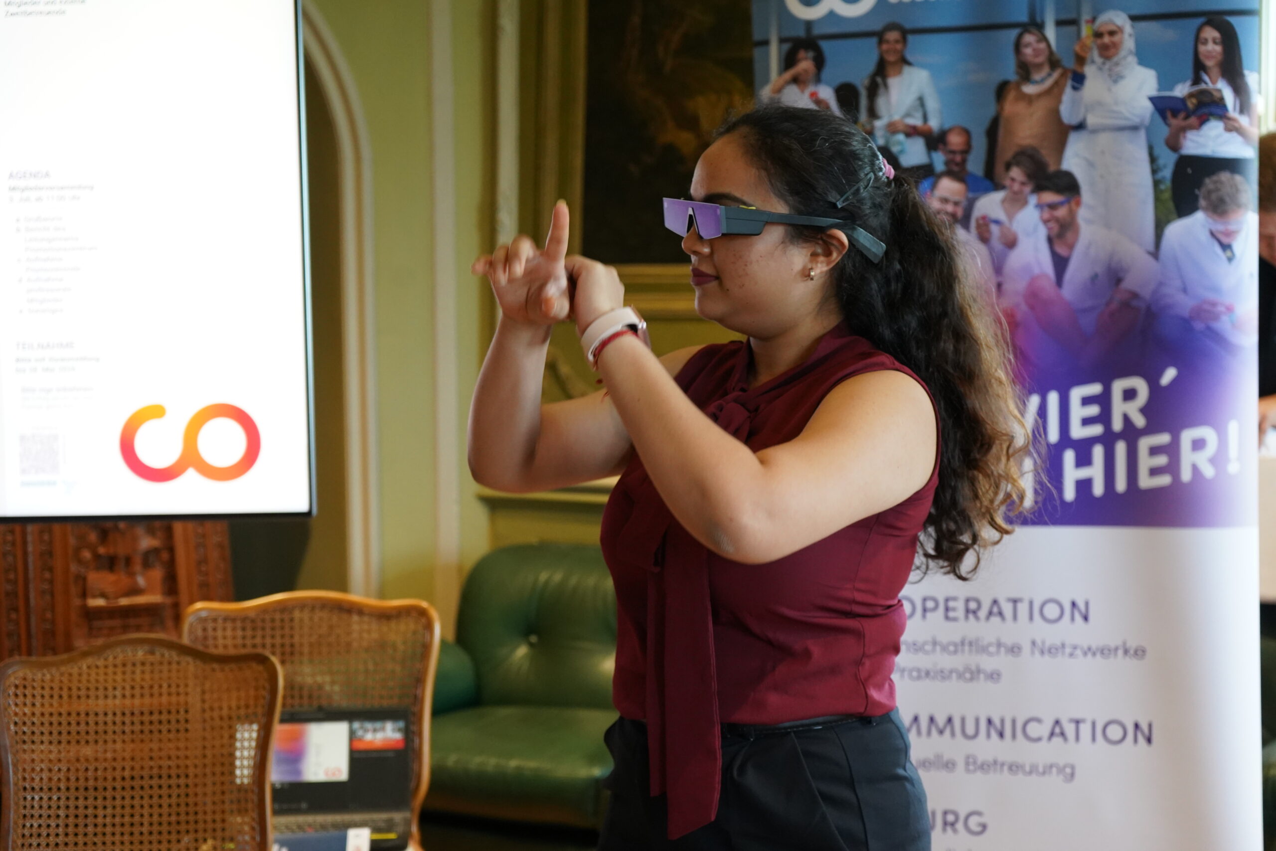 Eine Frau mit dunkler Sonnenbrille kommuniziert vor einer Gruppe in Gebärdensprache. Sie steht in einem Raum in der Nähe eines Banners und eines grünen Sofas und nimmt an einer offenbar formellen Promotionstagung oder einem Workshop teil, bei dem der Austausch und die Vorstellung guter Doktorarbeiten im Mittelpunkt stehen. at Hochschule Coburg