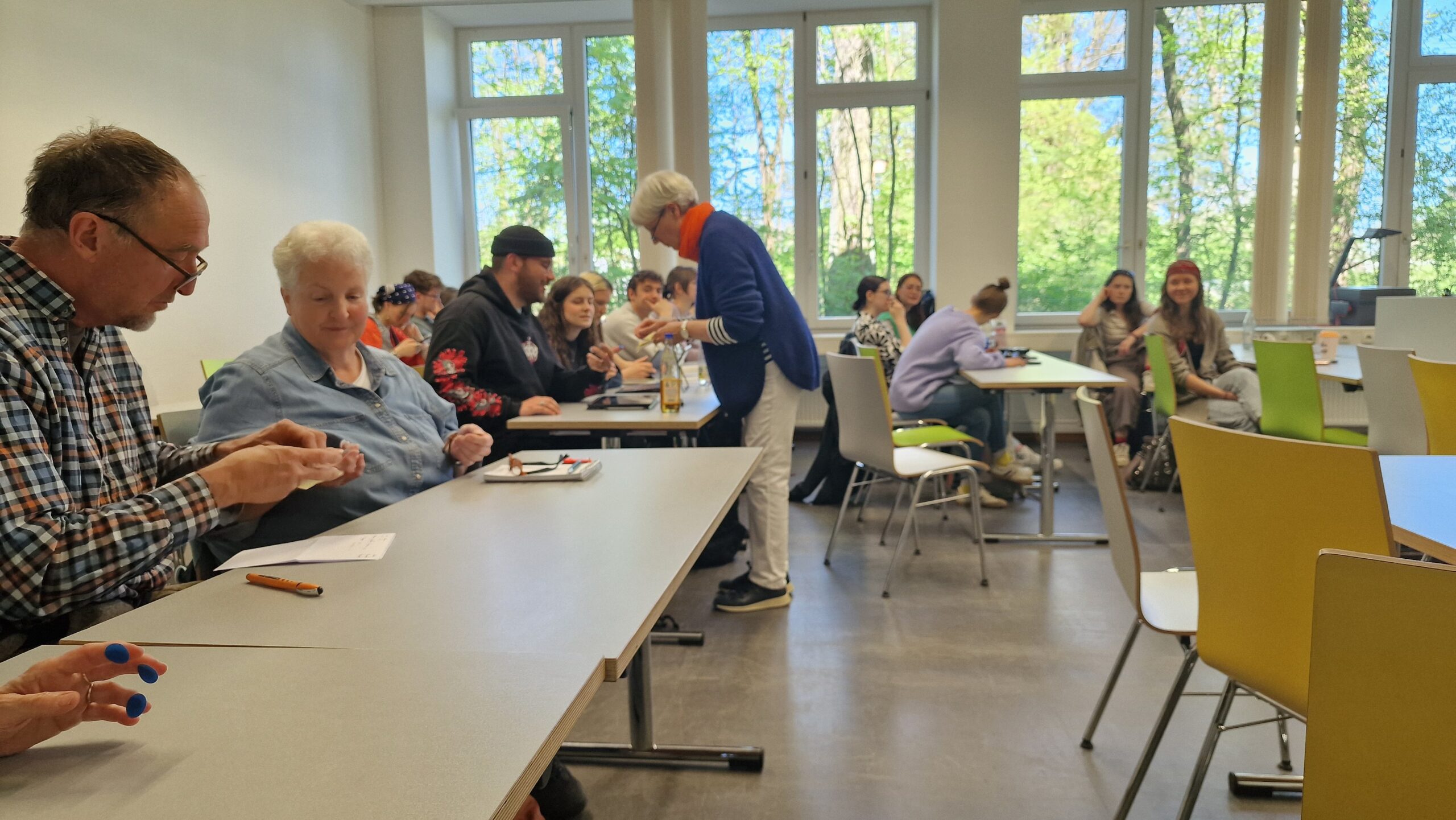 Eine Gruppe von Menschen sitzt an Tischen in einem hellen Klassenzimmer mit großen Fenstern, hinter denen Bäume zu sehen sind. Einige unterhalten sich, während eine Frau steht und zwischen den Tischen hin und her geht. at Hochschule Coburg