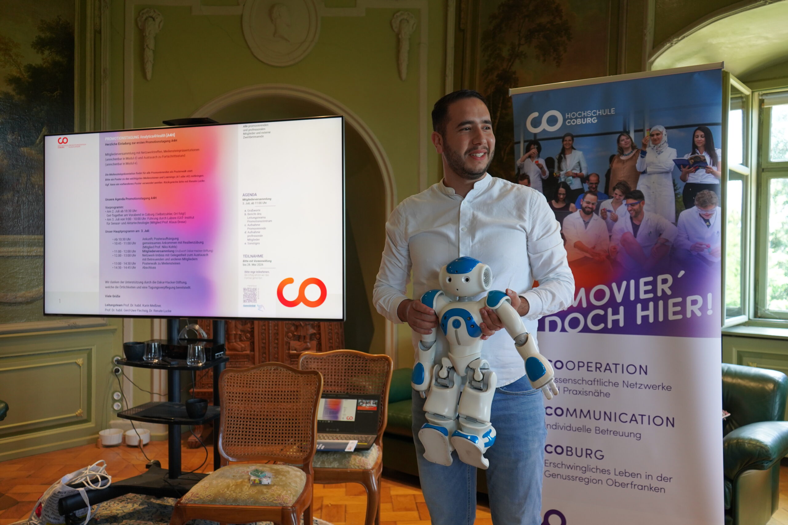 Ein Mann in einem weißen Hemd hält einen blau-weißen Roboter in einem Raum mit kunstvoll verzierten Wänden in der Hand, neben einem Fernseher, auf dem Text und ein Werbebanner für die Hochschule Coburg zu sehen sind, was eine inspirierende Atmosphäre für den Austausch und die Würdigung guter Doktorarbeiten während der Promotionstagung schafft. at Hochschule Coburg