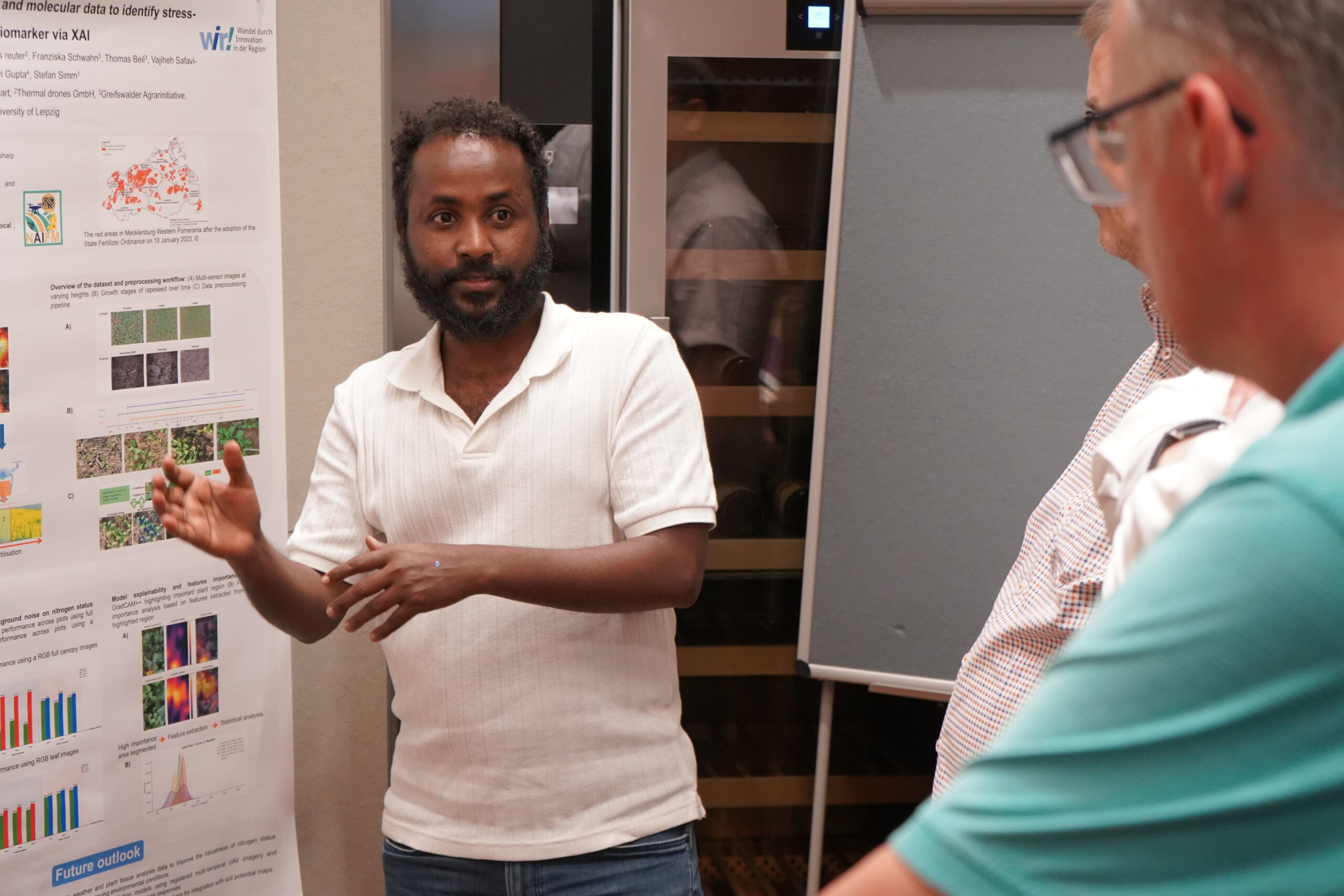 Ein Mann in einem weißen Hemd gestikuliert, während er zwei anderen Männern im Rahmen eines informellen Austauschs – vermutlich bei einer Promotionstagung oder einer wissenschaftlichen Veranstaltung zum Thema „gute Doktorarbeiten“ – seine Forschungsergebnisse auf einem Poster vorstellt. at Hochschule Coburg