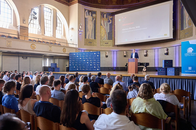 Ein Redner steht auf einem Podium auf der Bühne eines großen Saals und spricht zu den sitzenden Zuhörern. Hinter dem Redner befinden sich eine große Leinwand, auf der ein „Automatisch gespeicherter Entwurf“ zu sehen ist, sowie Banner; kunstvolle Verzierungen und hohe Fenster schmücken den Saal. at Hochschule Coburg