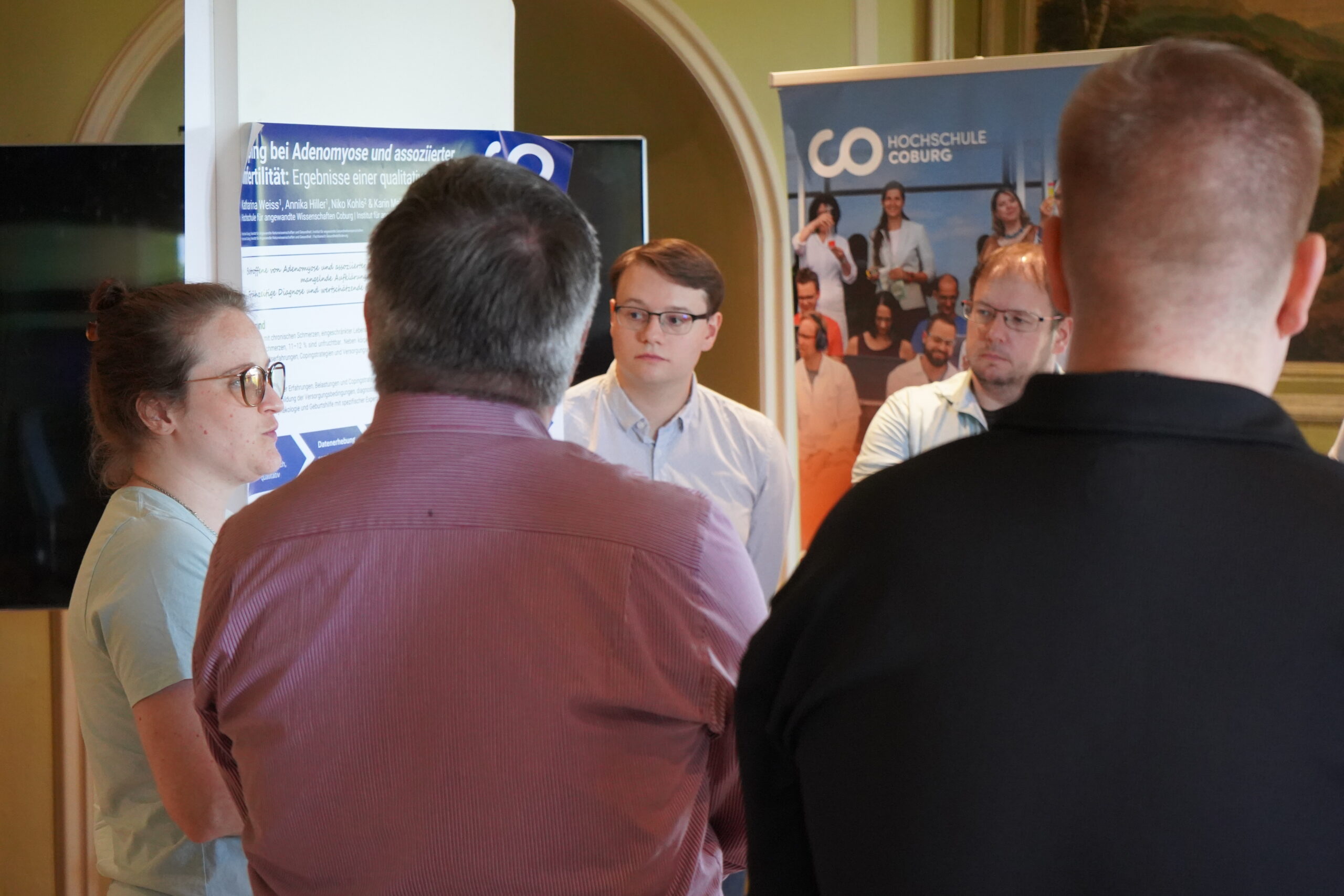 Fünf Personen stehen bei einer Promotionstagung in der Nähe von Plakaten und Bannern und unterhalten sich; zwei von ihnen blicken in die Kamera, drei stehen mit dem Rücken zur Kamera. Die Veranstaltung findet in einem professionellen Innenraum statt, der den Austausch fördert und gute Doktorarbeiten präsentiert. at Hochschule Coburg