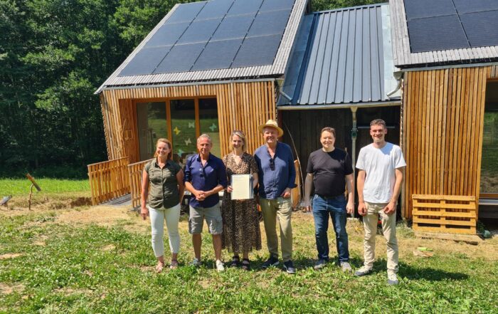 Sechs Menschen stehen lächelnd vor einem Holzhaus mit Sonnenkollektoren auf dem Dach. Eine Person hält eine Urkunde in der Hand, mit der ihre Bemühungen im Bereich „KlimaKulturKompetenz“ gewürdigt werden. Das Haus, das möglicherweise zum Waldkindergarten Pinzberg gehört, ist von Grünflächen umgeben, und im Hintergrund sind Bäume zu sehen. at Hochschule Coburg