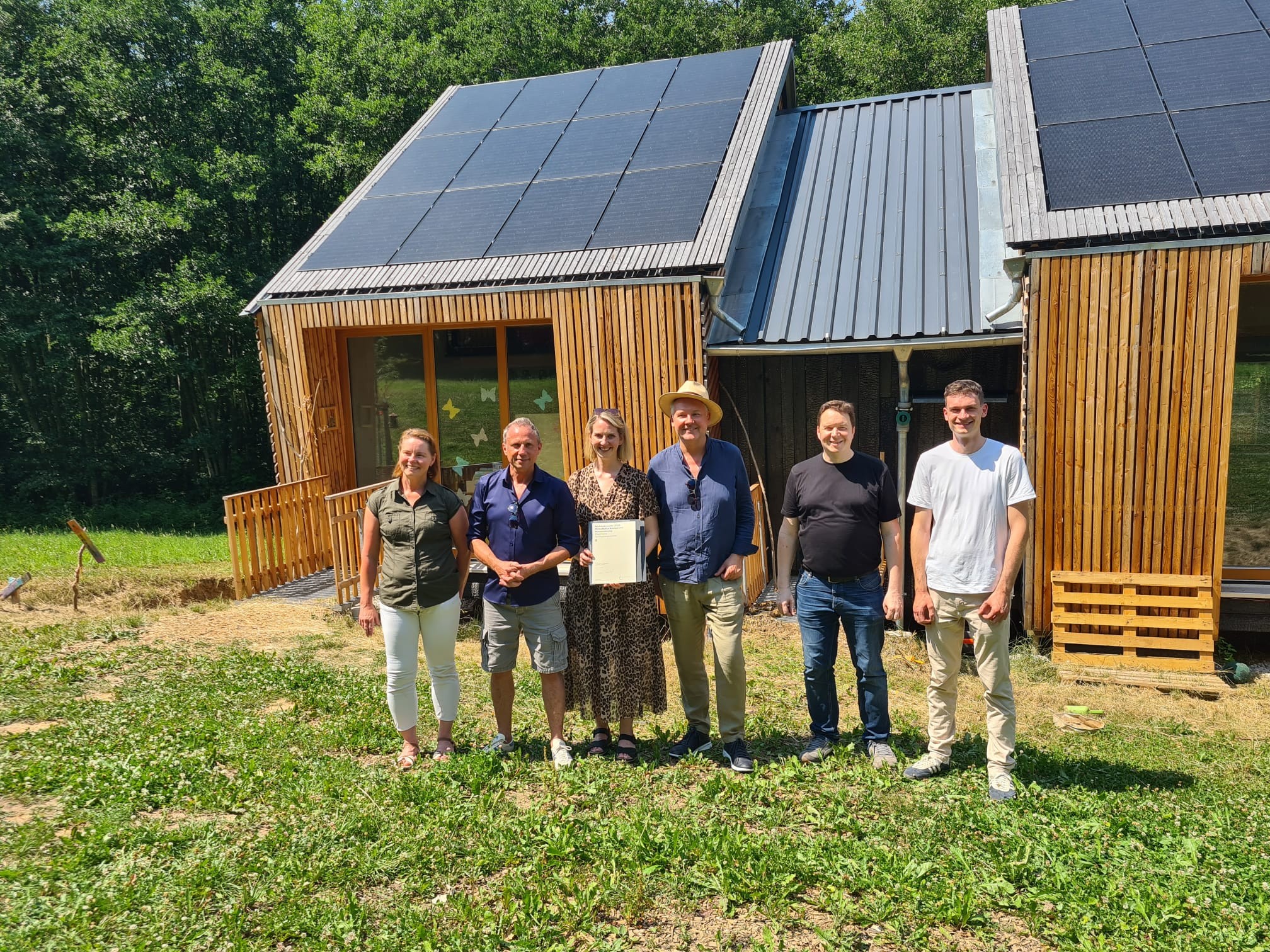 Sechs Menschen stehen lächelnd vor einem Holzhaus mit Sonnenkollektoren auf dem Dach. Eine Person hält eine Urkunde in der Hand, mit der ihre Bemühungen im Bereich „KlimaKulturKompetenz“ gewürdigt werden. Das Haus, das möglicherweise zum Waldkindergarten Pinzberg gehört, ist von Grünflächen umgeben, und im Hintergrund sind Bäume zu sehen. at Hochschule Coburg