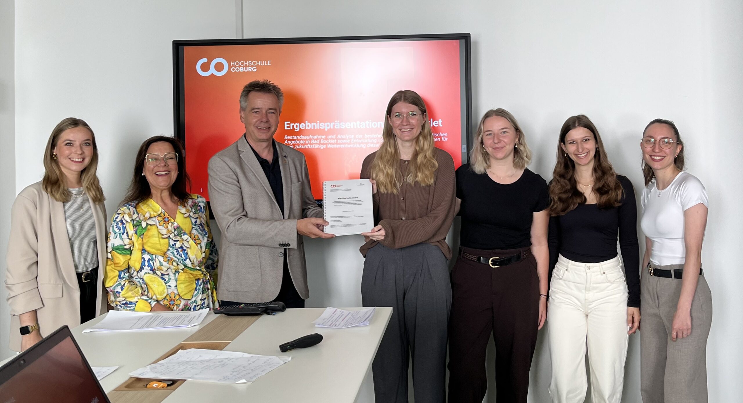 Sieben Personen stehen lächelnd in einem Konferenzraum und halten gemeinsam ein Dokument in den Händen. Auf einer Leinwand hinter ihnen ist eine rote Folie mit Text zu sehen. Auf dem Tisch vor ihnen liegen Unterlagen und ein Laptop. at Hochschule Coburg