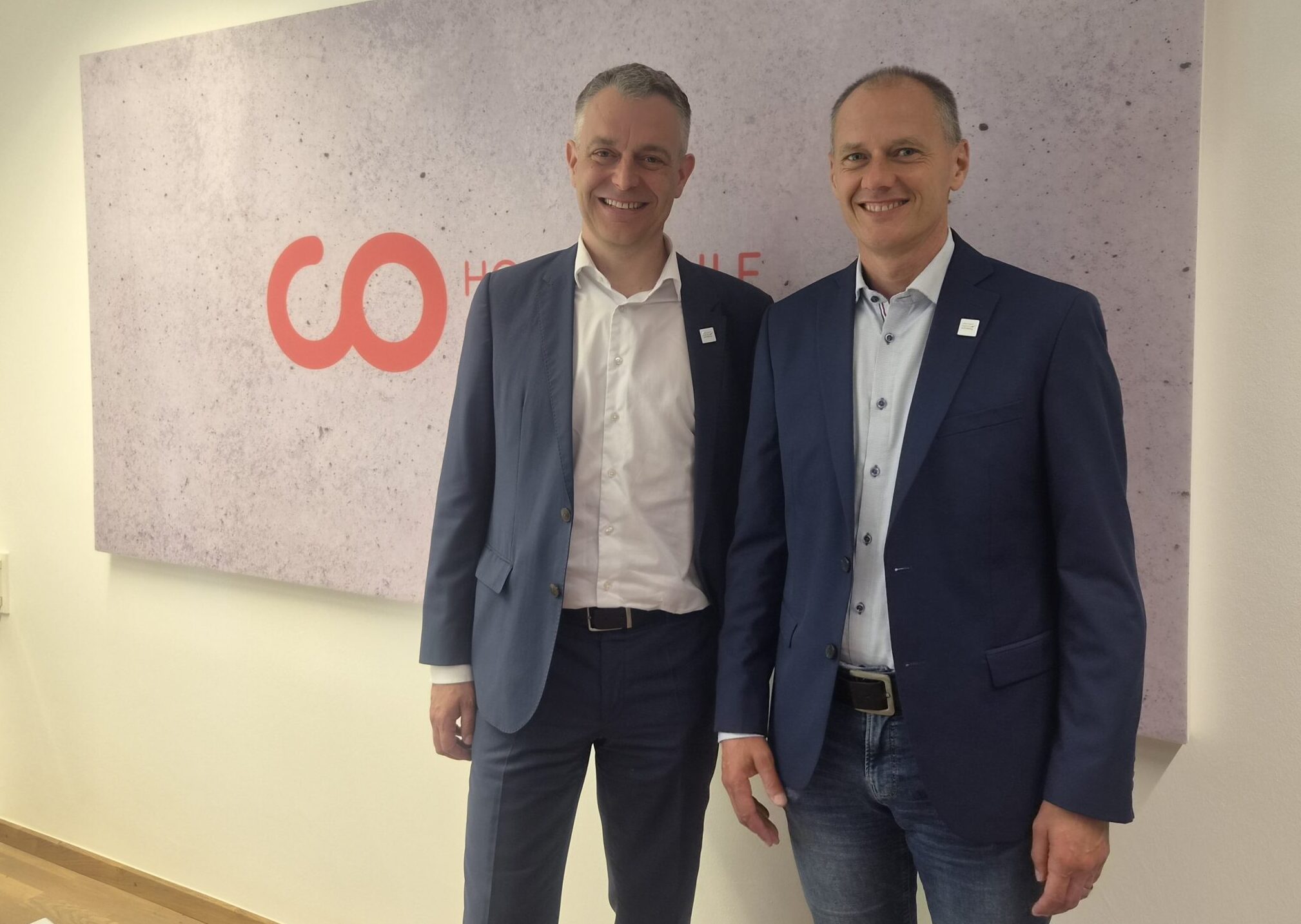 Zwei Männer in blauen Anzügen stehen lächelnd vor einem Wandschild mit roten „CO“-Buchstaben und Schriftzug in einem modernen Büroraum mit Holzboden – was die professionelle Atmosphäre widerspiegelt, die in Hochschulumgebungen innerhalb der Metropolregion häufig anzutreffen ist. at Hochschule Coburg