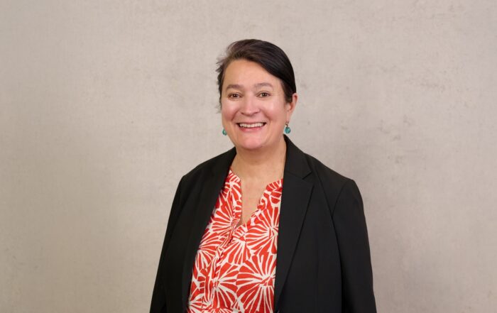 Eine Frau mit dunklem Haar, die einen schwarzen Blazer über einer rot-weiß gemusterten Bluse trägt, lächelt vor einer schlichten, hellen Wand – ihr selbstbewusstes Auftreten spiegelt ihr Engagement für den Katastrophenschutz und den sozialen Katastrophenschutz wider. at Hochschule Coburg