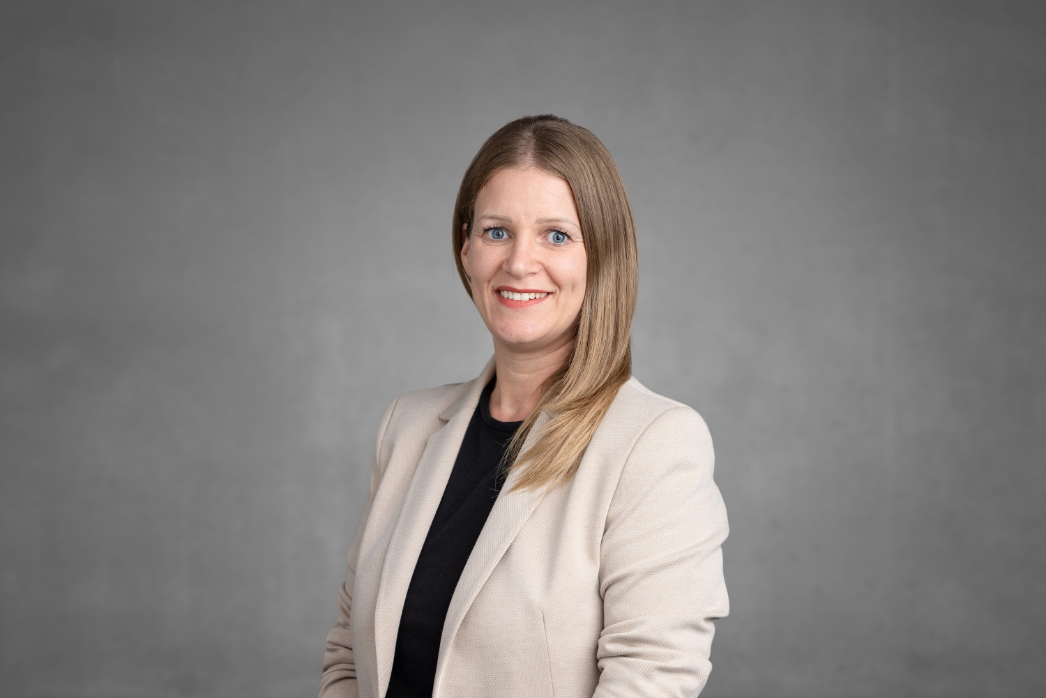 Eine Frau mit langen blonden Haaren, die einen beigen Blazer über einem schwarzen Oberteil trägt, steht lächelnd vor einem schlichten grauen Hintergrund und verkörpert die Professionalität und das Engagement, die im Bereich des Katastrophenschutzes oft zu beobachten sind. at Hochschule Coburg