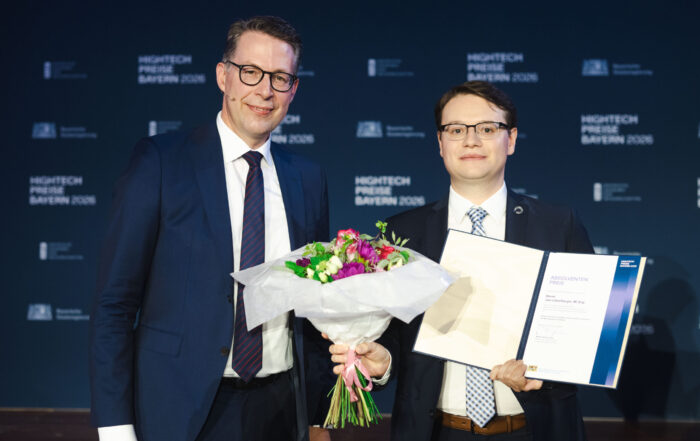 Zwei Männer in Anzügen posieren. Einer hält einen Blumenstrauß und eine Urkunde in der Hand. Beide lächeln. Im Hintergrund sind die Aufschrift „HIGHTECH PREIS BAYERN 2023“ sowie Logos in weißer Schrift auf blauem Hintergrund zu sehen, die die beim Hightech-Absolventenpreis Bayern gewürdigten Leistungen hervorheben und herausragende Coburger Nachwuchsforscher ehren. at Hochschule Coburg