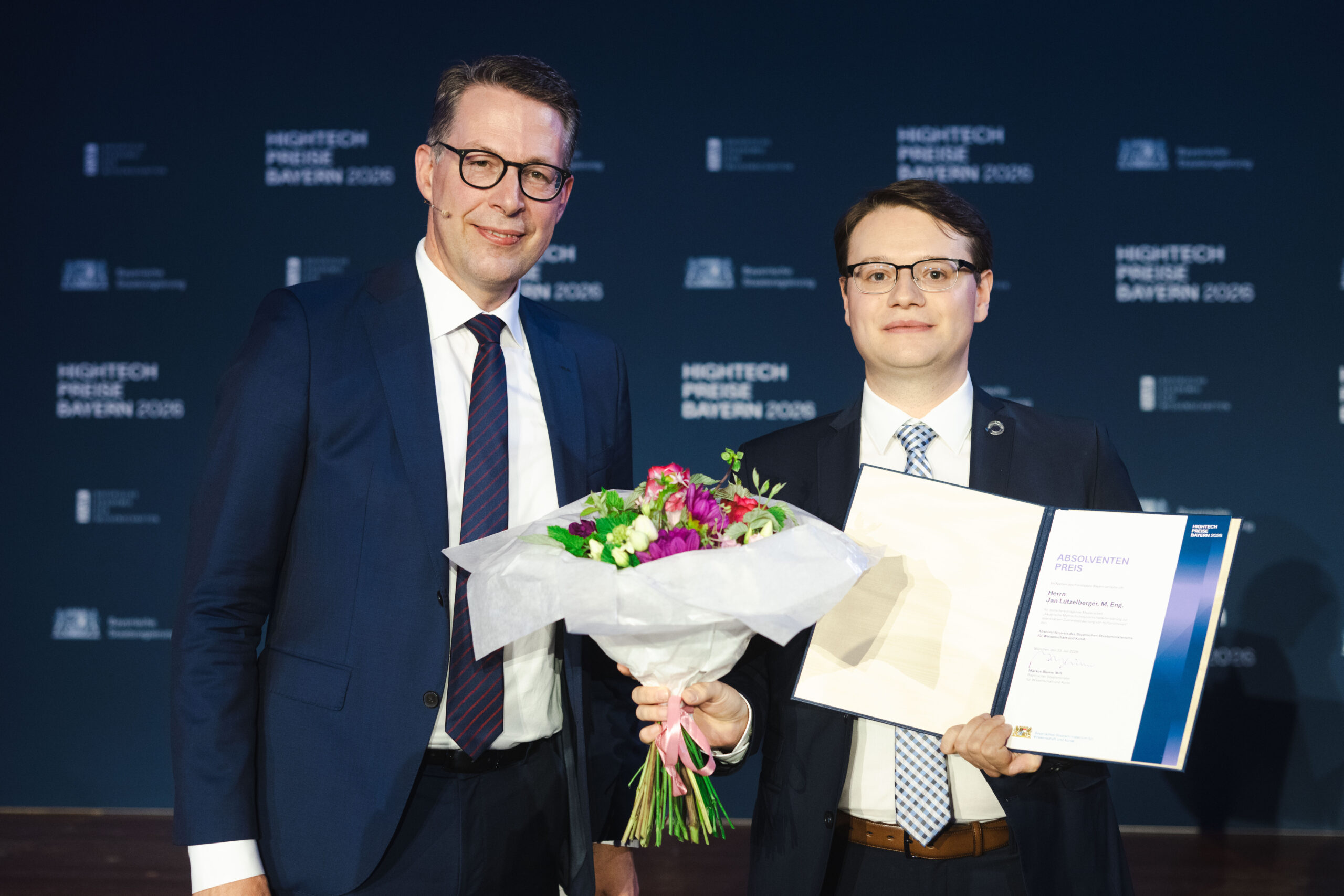 Zwei Männer in Anzügen posieren. Einer hält einen Blumenstrauß und eine Urkunde in der Hand. Beide lächeln. Im Hintergrund sind die Aufschrift „HIGHTECH PREIS BAYERN 2023“ sowie Logos in weißer Schrift auf blauem Hintergrund zu sehen, die die beim Hightech-Absolventenpreis Bayern gewürdigten Leistungen hervorheben und herausragende Coburger Nachwuchsforscher ehren. at Hochschule Coburg