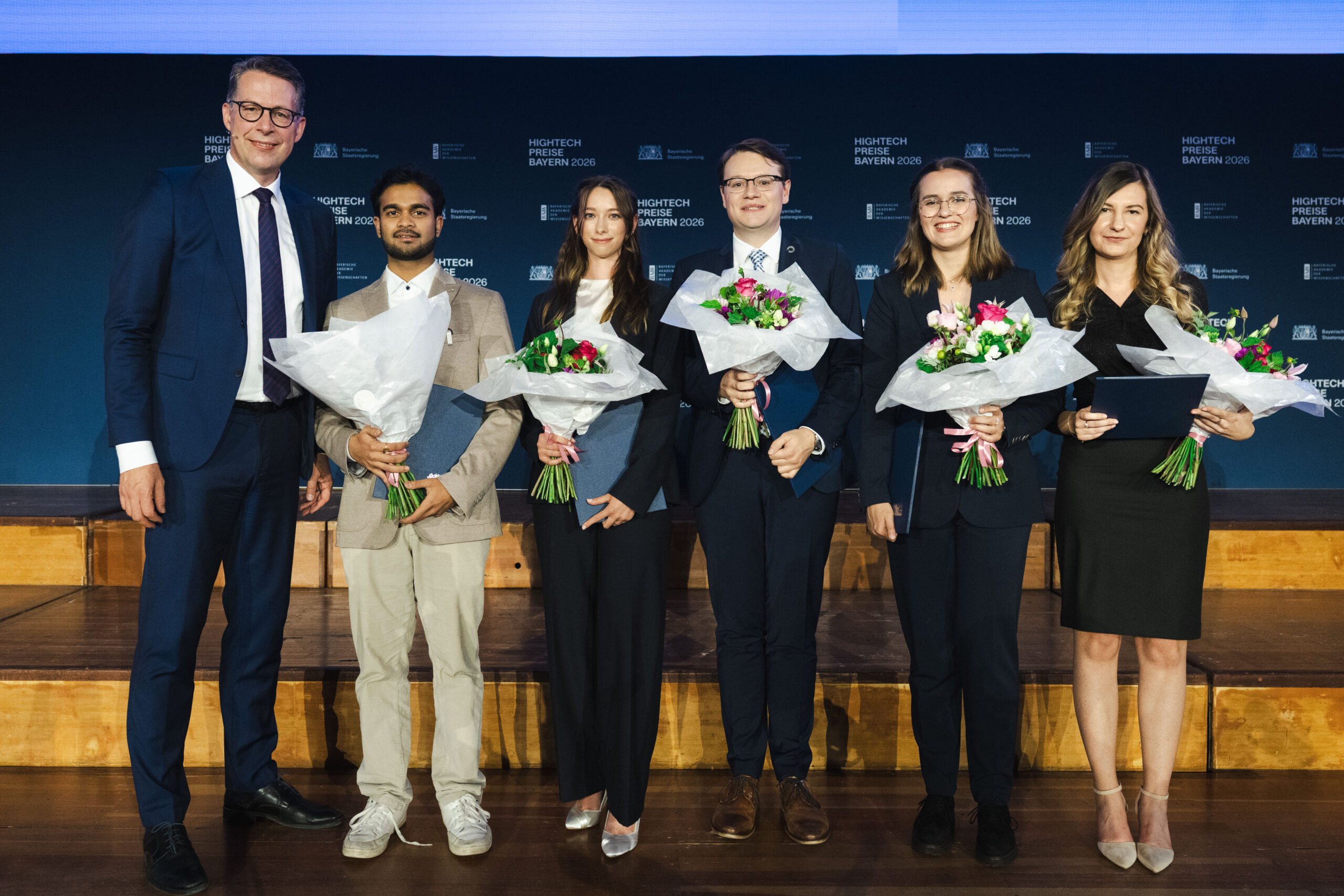 Sechs Personen in festlicher Kleidung stehen auf einer Bühne, fünf von ihnen halten Blumensträuße und Urkunden in den Händen und lächeln für ein Gruppenfoto. Hinter ihnen befindet sich ein blauer Hintergrund mit weißer Schrift, auf dem der „Hightech-Absolventenpreis Bayern“ hervorgehoben ist. Bei der Veranstaltung werden herausragende Nachwuchsforscher aus Coburg für ihre Leistungen geehrt. at Hochschule Coburg