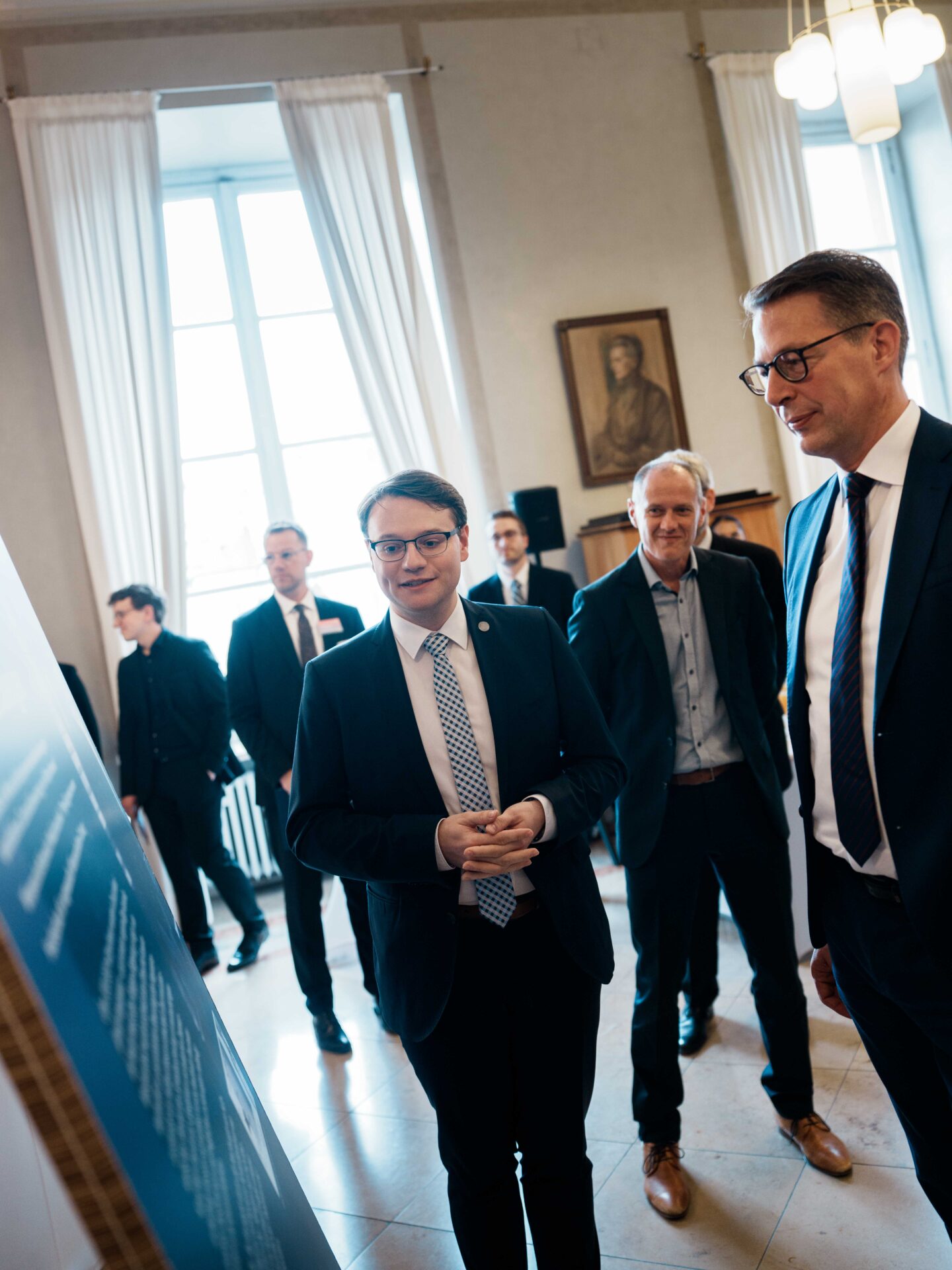 Eine Gruppe von Männern in Anzügen steht im Innenraum und betrachtet aufmerksam eine Präsentationswand während der Veranstaltung „Coburger Nachwuchsforscher“. Der Raum verfügt über große Fenster mit weißen Vorhängen und ein Porträt an der Wand im Hintergrund, was eine elegante Atmosphäre für die Verleihung des „Hightech-Absolventenpreises Bayern“ schafft. at Hochschule Coburg