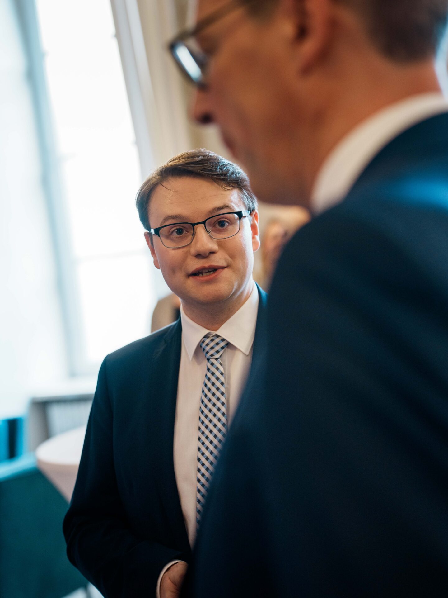 Ein Mann im Anzug und mit Brille, möglicherweise ein Preisträger des „Hightech-Absolventenpreises Bayern“, unterhält sich mit einem anderen Mann im Anzug, der im Vordergrund unscharf zu sehen ist. Sie befinden sich in einem Raum in der Nähe eines Fensters, durch das Tageslicht fällt, und sprechen möglicherweise über Themen im Zusammenhang mit der Initiative „Coburger Nachwuchsforscher“. at Hochschule Coburg