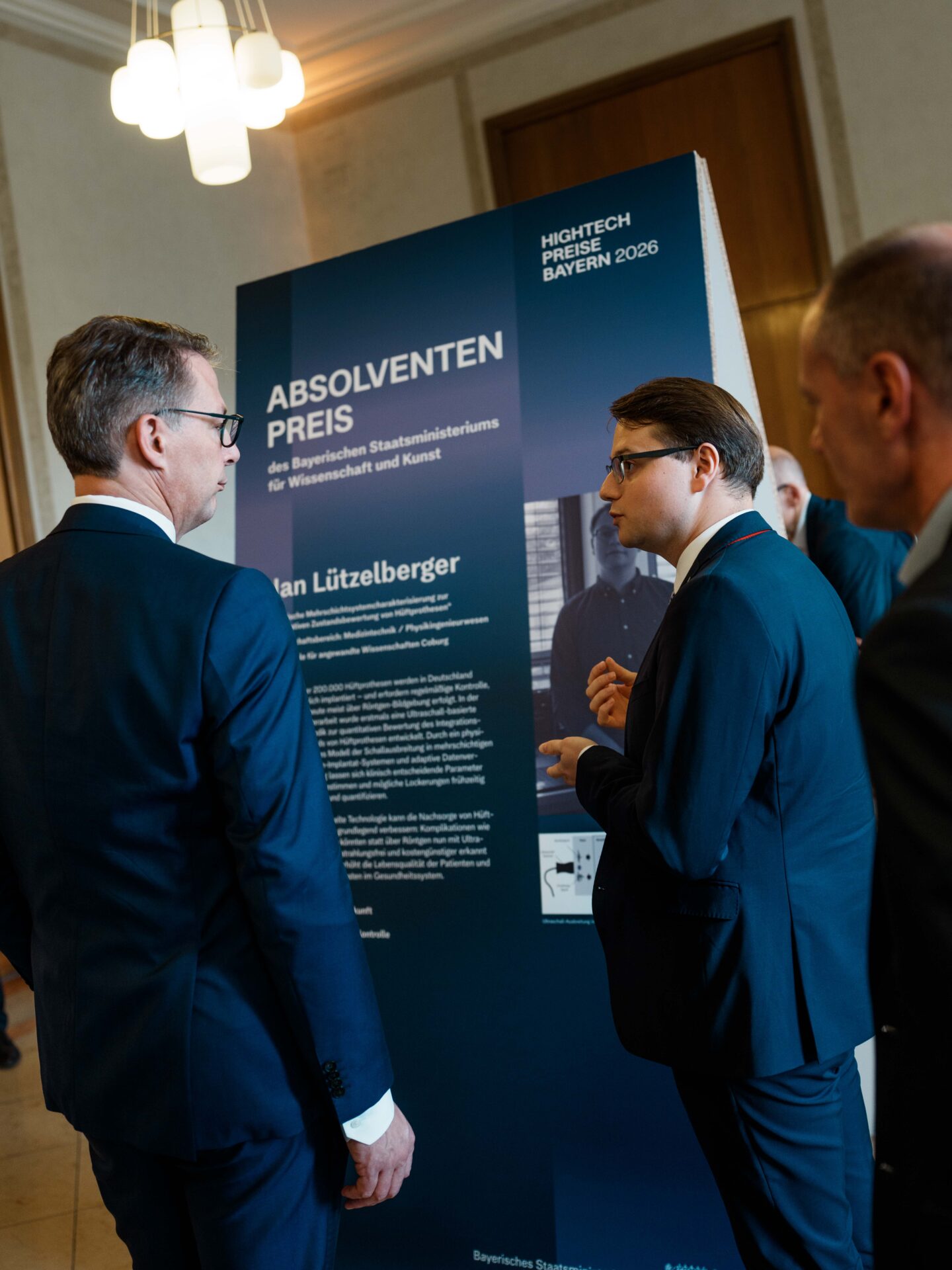 Drei Männer in Anzügen stehen bei einer feierlichen Veranstaltung in einem Saal neben einer großen Anschlagtafel zum „Absolventenpreis“. Die Tafel, auf der Leistungen wie der „Hightech-Absolventenpreis Bayern“ hervorgehoben werden, enthält einen Text und ein Schwarz-Weiß-Foto. Der Anlass scheint eine Konferenz oder eine Feier zu Ehren herausragender Coburger Nachwuchsforscher zu sein. at Hochschule Coburg