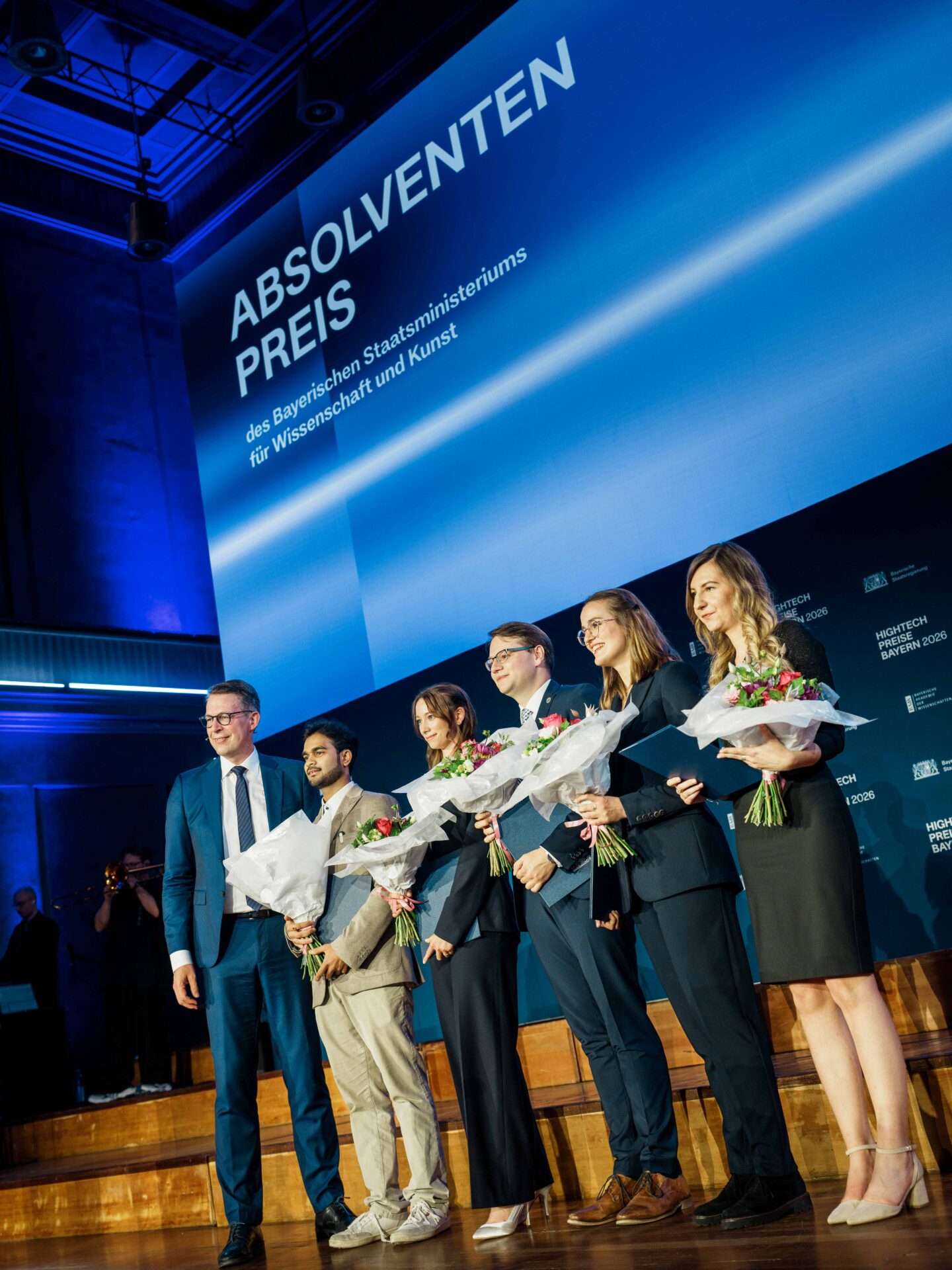 Fünf Preisträger mit Blumen in den Händen stehen bei einer Feier in einem Saal auf der Bühne neben einem Mann im Anzug unter einem blauen Schild mit der Aufschrift „Absolventenpreis“ und feiern die Leistungen der Coburger Nachwuchsforscher im Rahmen des renommierten Hightech-Absolventenpreises Bayern. at Hochschule Coburg