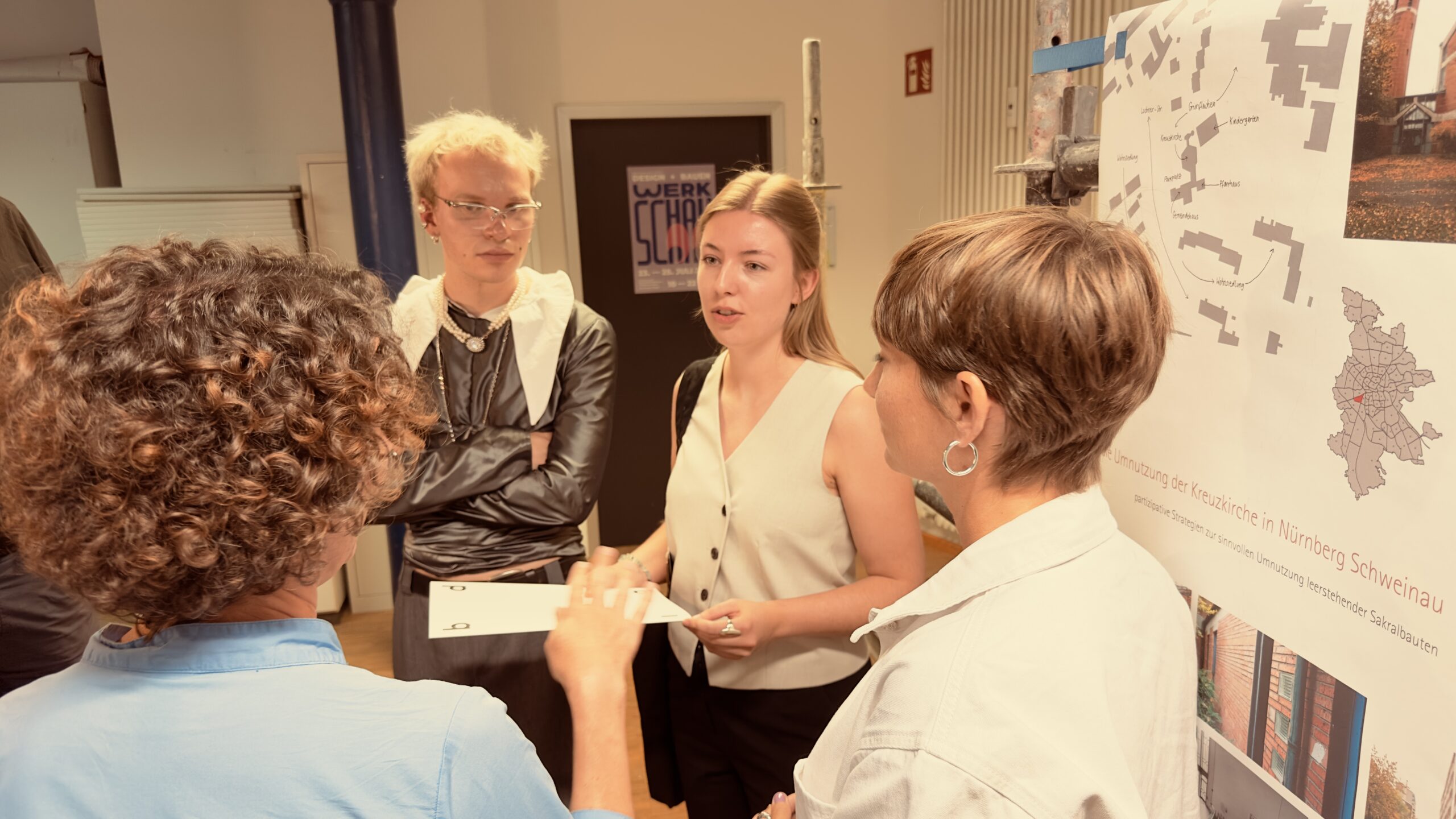 Vier Personen stehen in einer kleinen Gruppe beieinander und unterhalten sich neben einer Wand, an der eine Karte und Unterlagen angebracht sind; möglicherweise besprechen sie Projekteinreichungen für die bdia-Preisverleihung. Eine Person gestikuliert, während die anderen aufmerksam zuhören, darunter zwei Studentinnen, die kürzlich für ihre innovativen Ideen ausgezeichnet wurden. Der Schauplatz scheint ein Arbeitsbereich oder ein Besprechungsraum zu sein. at Hochschule Coburg