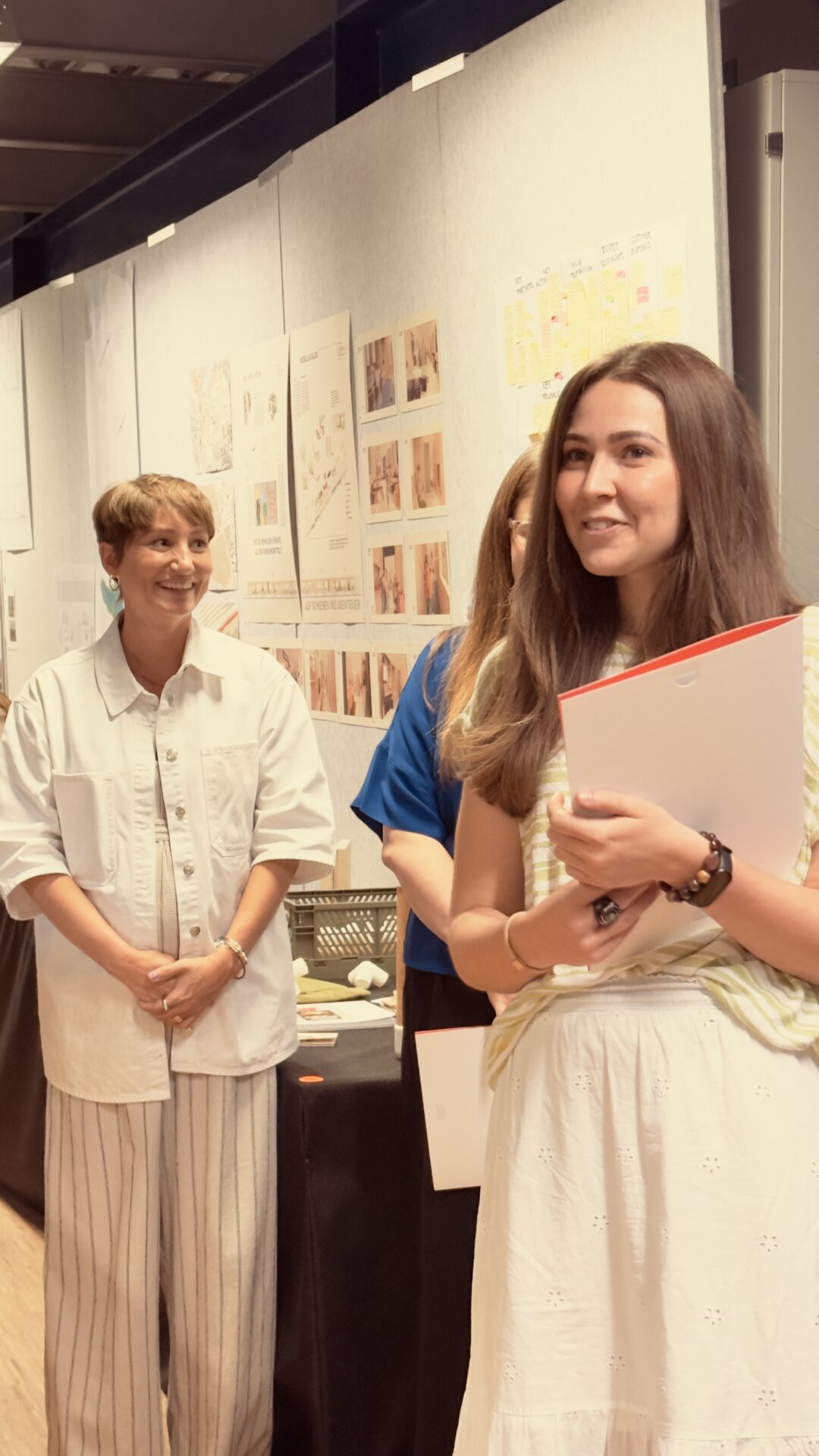Vor einer Ausstellung mit Skizzen und Diagrammen standen drei Studentinnen, die ausgezeichnet wurden. Eine Frau im Vordergrund hält eine Mappe in der Hand und lächelt, während die anderen hinter ihr stehen und ebenfalls lächeln. Sie scheinen sich auf einer Preisverleihung oder einer Ausstellung zu befinden. at Hochschule Coburg