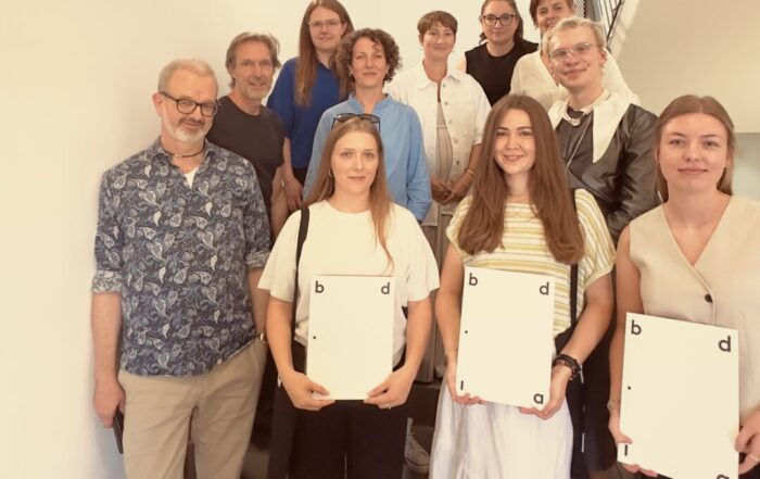 Eine Gruppe von elf Personen steht auf einer Treppe, wobei vier Frauen ganz vorne ihre Urkunden in den Händen halten – ein klarer Höhepunkt der bdia-Preisverleihung. Alle lächeln und blicken in die Kamera, während sie sich in einem gut beleuchteten Innenraum befinden; die für ihre Leistungen ausgezeichneten Studentinnen präsentieren stolz ihre Auszeichnungen. at Hochschule Coburg