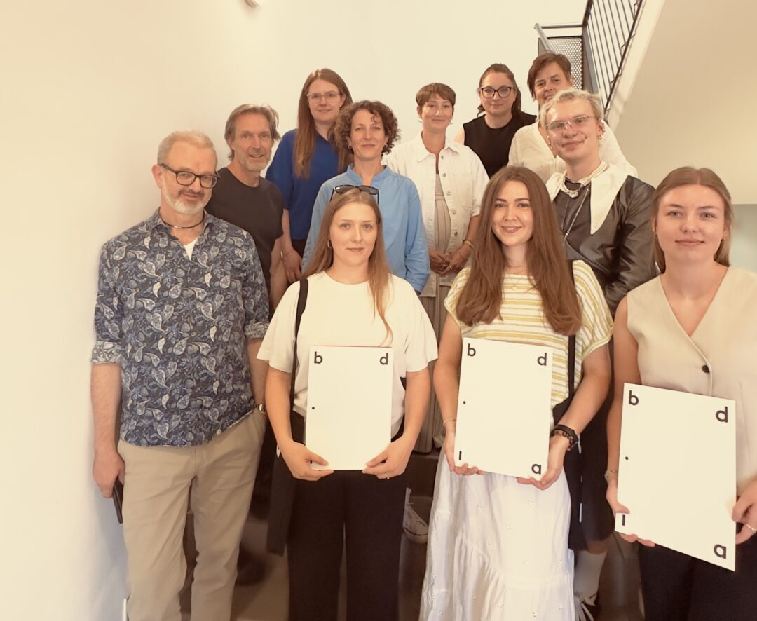 Eine Gruppe von elf Personen steht auf einer Treppe, wobei vier Frauen ganz vorne ihre Urkunden in den Händen halten – ein klarer Höhepunkt der bdia-Preisverleihung. Alle lächeln und blicken in die Kamera, während sie sich in einem gut beleuchteten Innenraum befinden; die für ihre Leistungen ausgezeichneten Studentinnen präsentieren stolz ihre Auszeichnungen. at Hochschule Coburg