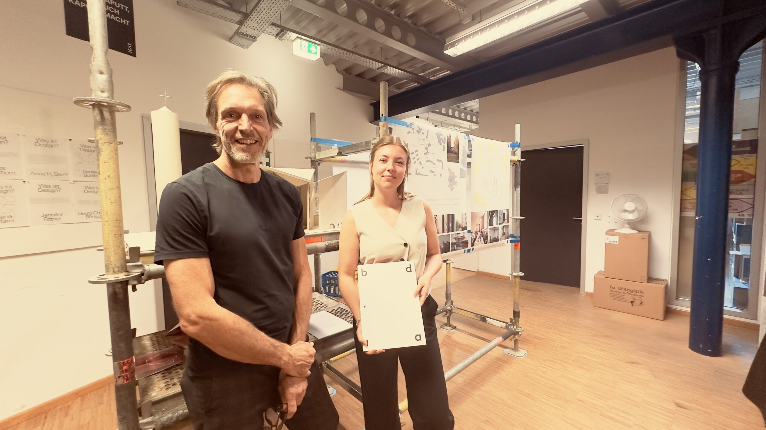 Zwei Studentinnen stehen in einem modernen Arbeitsbereich, hinter ihnen sind Baupläne und Tafeln zu sehen. Die Person auf der rechten Seite hält ein Dokument in der Hand – wahrscheinlich von der bdia-Preisverleihung – und beide lächeln in die Kamera. Die Umgebung wirkt kreativ und hell. at Hochschule Coburg