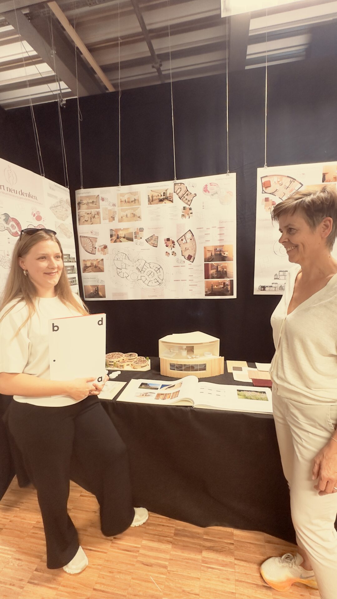 Zwei Frauen stehen neben einer Architekturausstellung, bei der Zeichnungen, Fotos und ein Gebäudemodell auf einem Tisch ausgestellt sind. Eine der Frauen, die kürzlich bei der Preisverleihung des bdia als Studentin ausgezeichnet wurde, hält Unterlagen in der Hand und lächelt, während die andere daneben steht. at Hochschule Coburg