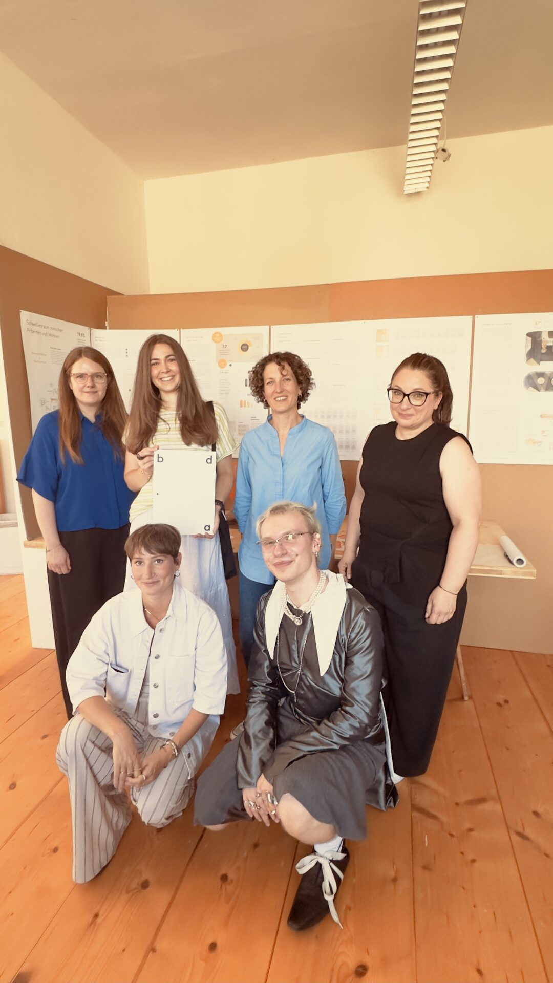 Sechs Personen posieren und lächeln in einem Raum mit Holzboden und Präsentationstafeln an der Wand. Eine Person steht da und hält ein Dokument in der Hand, das wahrscheinlich von der Preisverleihung des bdia stammt, bei der Studentinnen ausgezeichnet wurden. Der Rahmen scheint eine akademische oder berufliche Präsentation zu sein. at Hochschule Coburg