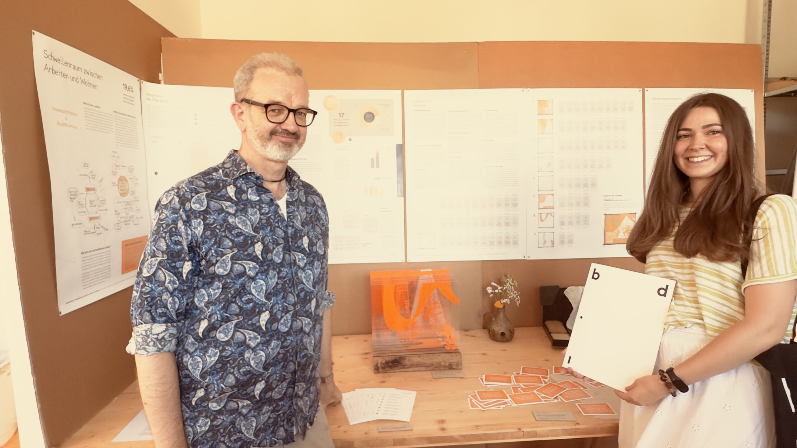Ein Mann und eine Frau stehen an einem Holztisch, auf dem Papiere und eine kleine orangefarbene Skulptur liegen, und lächeln vor Präsentationswänden mit Tabellen und Diagrammen bei der bdia-Preisverleihung, bei der Studentinnen ihre innovativen Projekte vorstellen. at Hochschule Coburg