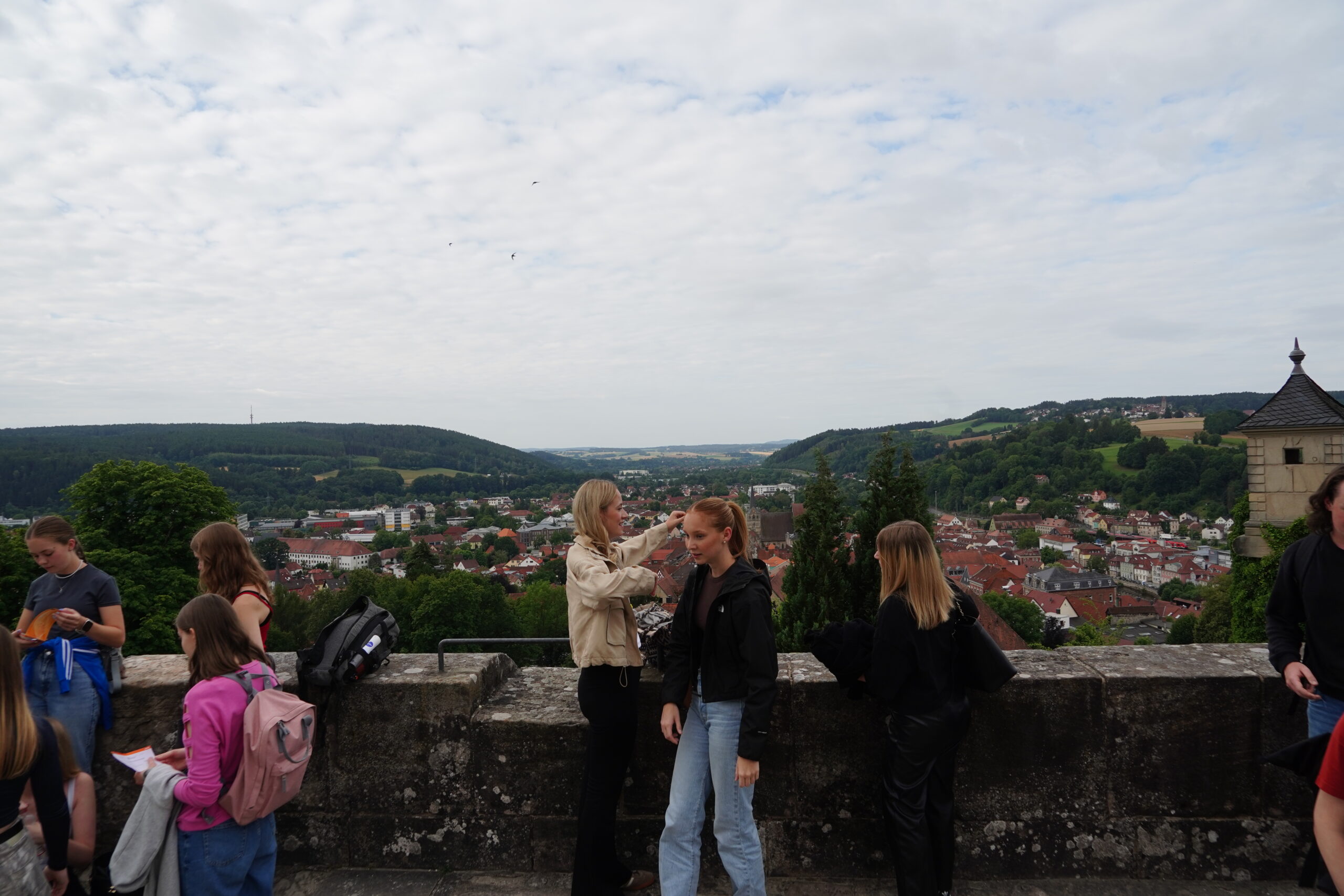 Mehrere Jugendliche stehen auf einer Steinterrasse und genießen den malerischen Ausblick auf Kronach – mit der charmanten Altstadt unter ihnen, den grünen Hügeln und dem bewölkten Himmel über ihnen. Einige unterhalten sich oder schauen auf ihre Handys; im Hintergrund erstrecken sich Bäume und Dächer, und so entsteht ein entspannter Moment, der vielleicht sogar Joey Kelly ins Auge gefallen wäre. at Hochschule Coburg