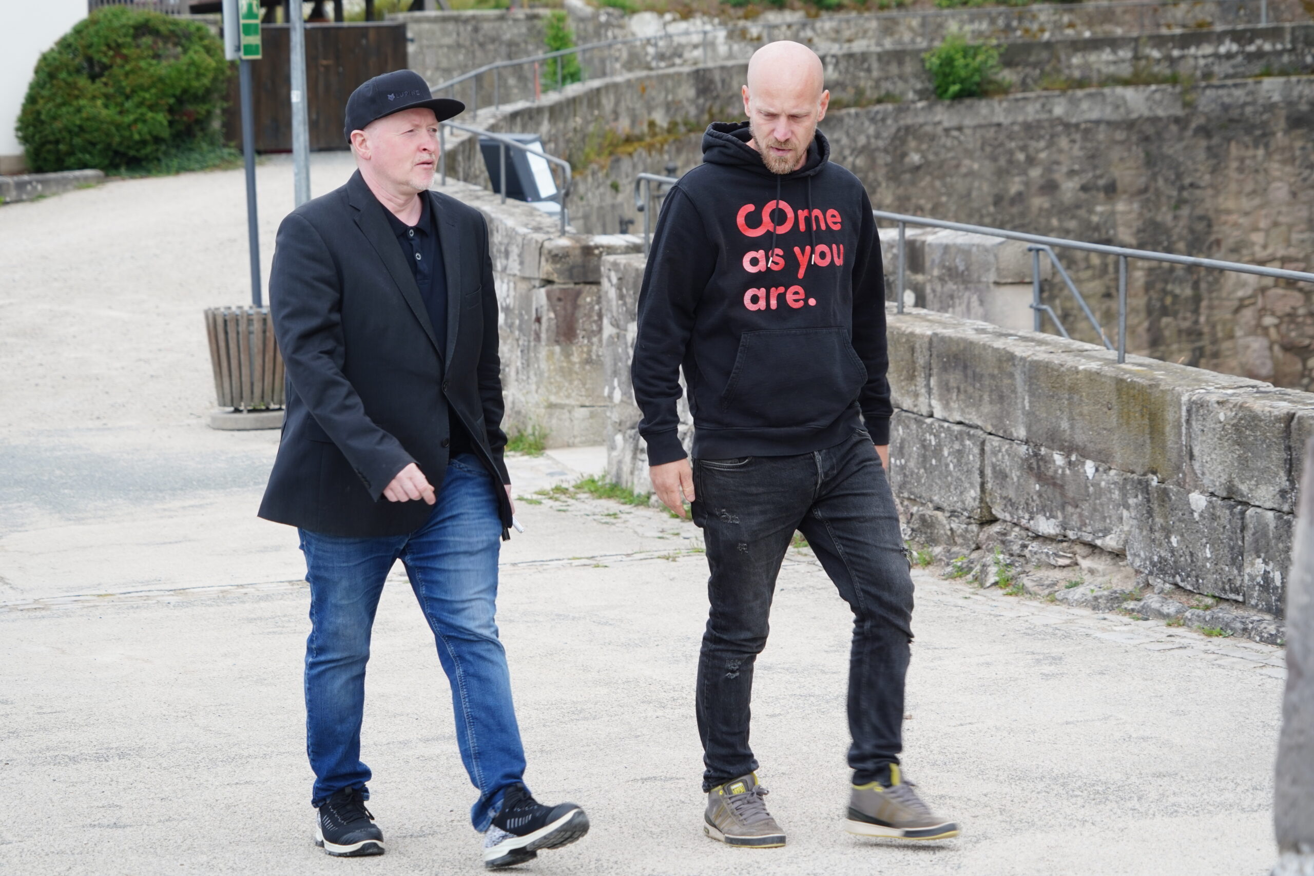 Zwei Männer, möglicherweise in Kronach, gehen draußen auf einem gepflasterten Weg in der Nähe einer Steinmauer spazieren; der eine trägt einen schwarzen Blazer und eine Mütze, während der andere einen schwarzen Kapuzenpulli mit der Aufschrift „Come as you are“ trägt. Ihr ungezwungener Spaziergang könnte Jugendliche, die zu Vorbildern wie Joey Kelly aufschauen, leicht inspirieren. at Hochschule Coburg
