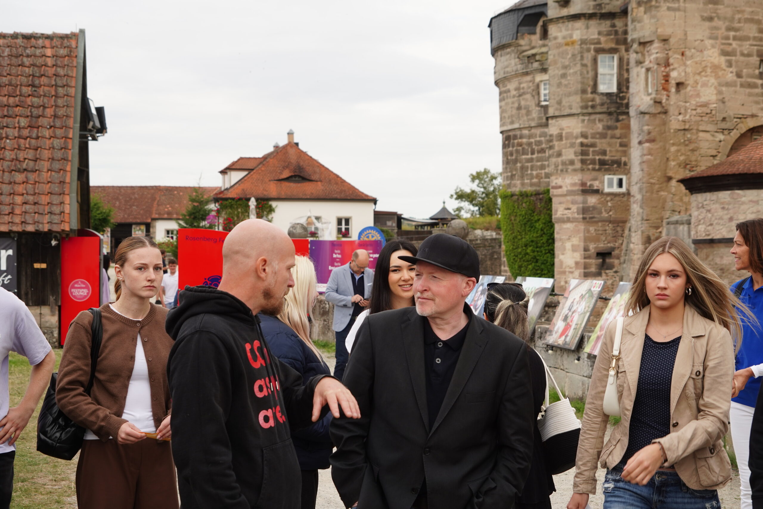 Menschen, darunter auch Jugendliche, spazieren vor historischen Steingebäuden und roten Verkaufsständen in Kronach entlang und unterhalten sich. Einige blicken direkt in die Kamera, während andere sich unterhalten oder lässig vorbeischlendern. Die Szene wirkt in dieser städtischen Umgebung im Freien entspannt – vielleicht wartet man sogar darauf, jemanden wie Joey Kelly zu erblicken. at Hochschule Coburg