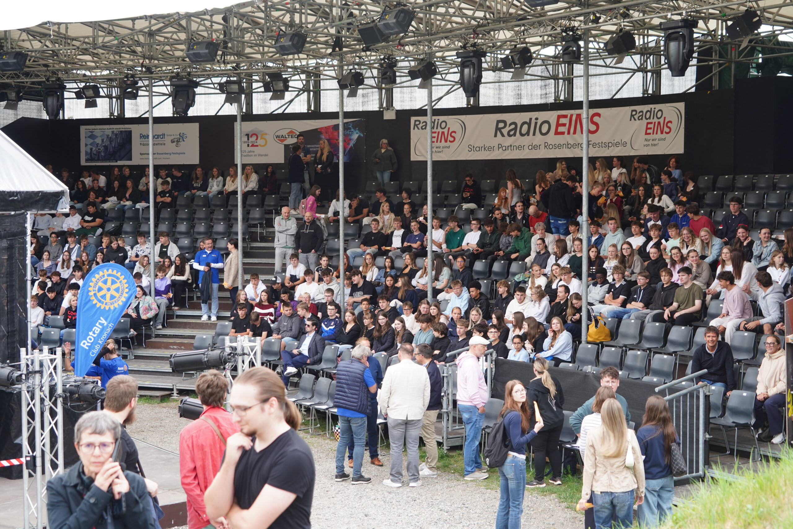 Eine große Menschenmenge, darunter viele Jugendliche, sitzt und steht in einer Freiluftarena in Kronach; einige unterhalten sich, andere schauen zu. Auf den Transparenten darüber sind RadioEINS und Rotary zu sehen. Der Bereich verfügt über gestufte Sitzplätze und eine Überdachung; als besonderer Gast wird Joey Kelly erwartet. at Hochschule Coburg
