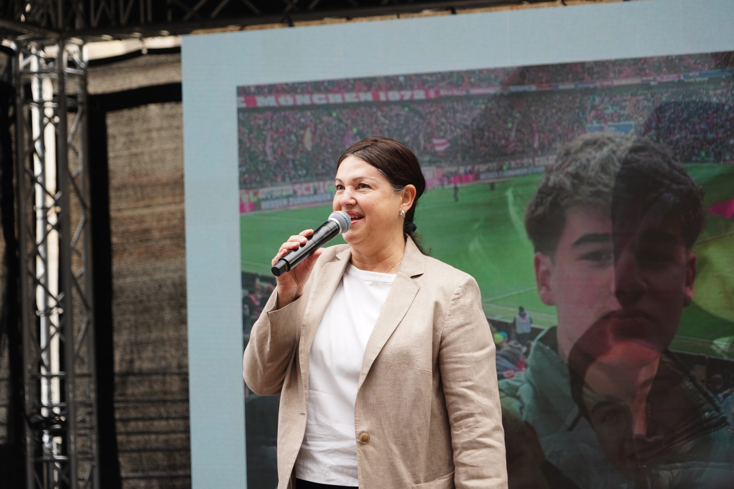 Eine Frau in einem beigen Blazer spricht auf der Bühne in Kronach in ein Mikrofon, während hinter ihr auf einer großen Leinwand das Publikum in einem Stadion und ein Teil des Gesichts eines jungen Mannes – möglicherweise Joey Kelly – zu sehen sind, was die Aufmerksamkeit der Jugendlichen im Publikum auf sich zieht. at Hochschule Coburg