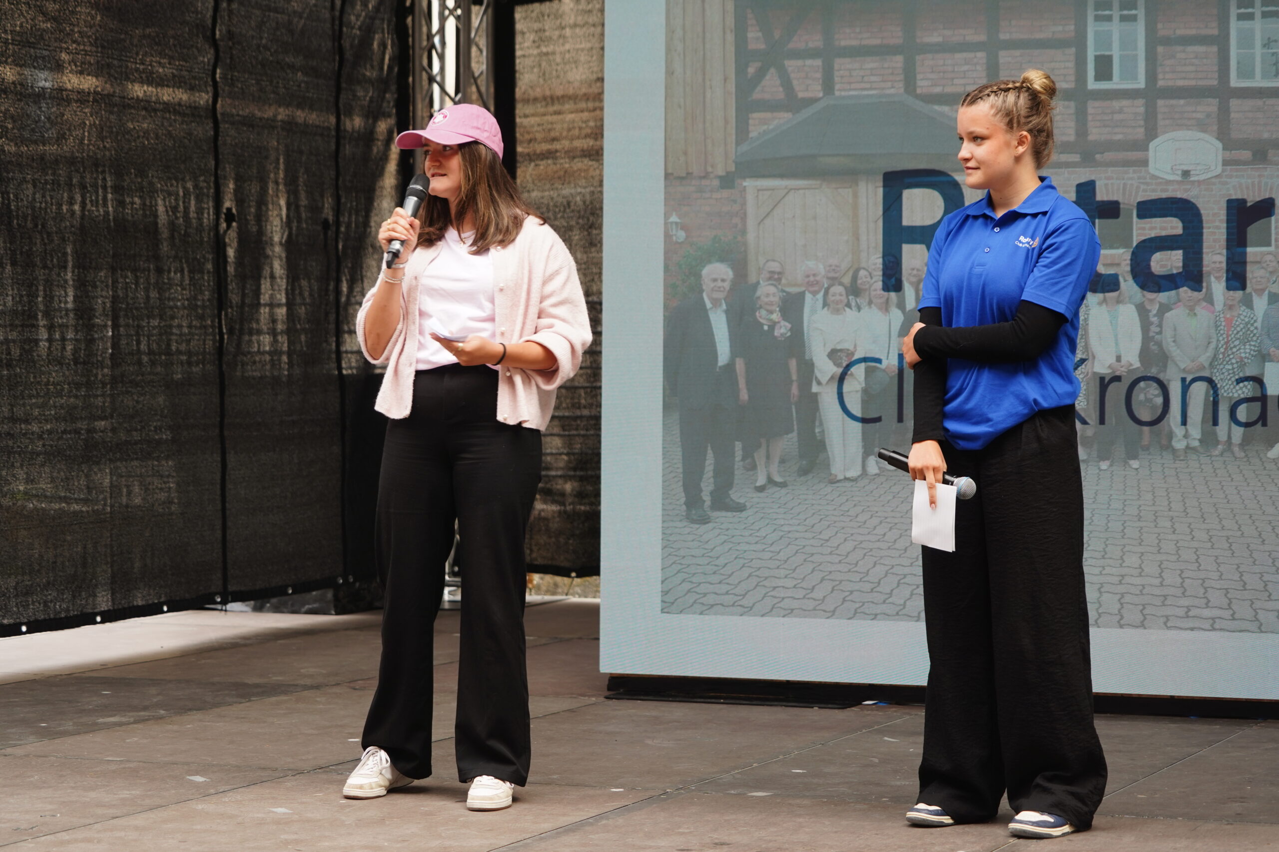 Auf der Bühne in Kronach stehen zwei Frauen; die eine in einer rosa Jacke spricht ins Mikrofon, während die andere in einem blauen Hemd Papiere in der Hand hält. Hinter ihnen sind ein projiziertes Bild – möglicherweise von Joey Kelly – und Text zu sehen, während sie sich an die anwesenden Jugendlichen wenden. at Hochschule Coburg