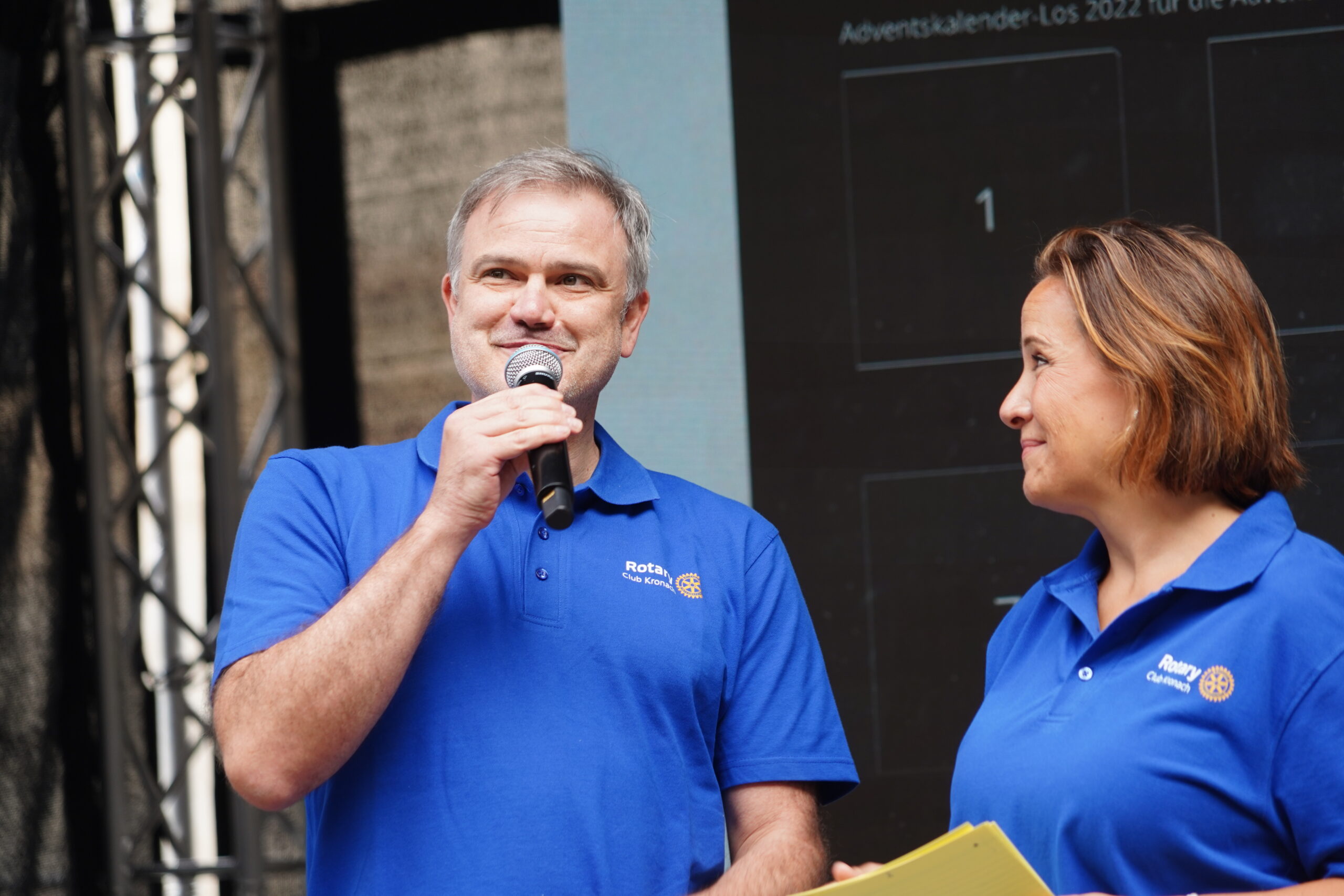 Zwei Personen in identischen blauen Rotary-Shirts stehen auf der Bühne in Kronach. Der Mann links lächelt und spricht in ein Mikrofon – vielleicht stellt er gerade den Ehrengast Joey Kelly vor –, während die Frau rechts ihn ansieht, einen gelben Notizblock in der Hand hält und mit den anwesenden Jugendlichen ins Gespräch kommt. at Hochschule Coburg