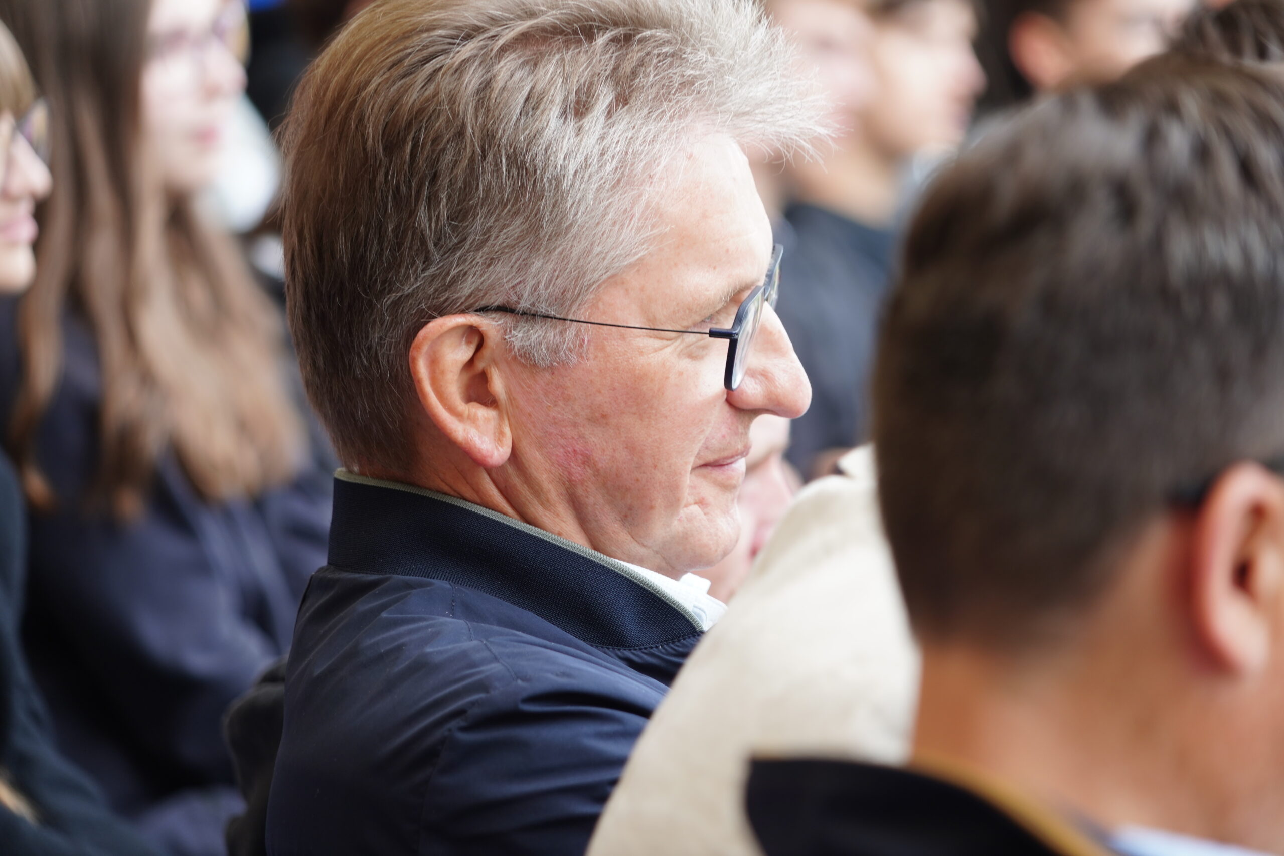 Ein Mann mittleren Alters mit Brille und grauen Haaren sitzt inmitten einer Gruppe von Menschen, blickt nach vorne und lächelt leicht. Der Schauplatz scheint eine Veranstaltung oder ein Treffen in Kronach zu sein, an der möglicherweise Jugendliche teilnehmen und bei der Joey Kelly auftritt. at Hochschule Coburg