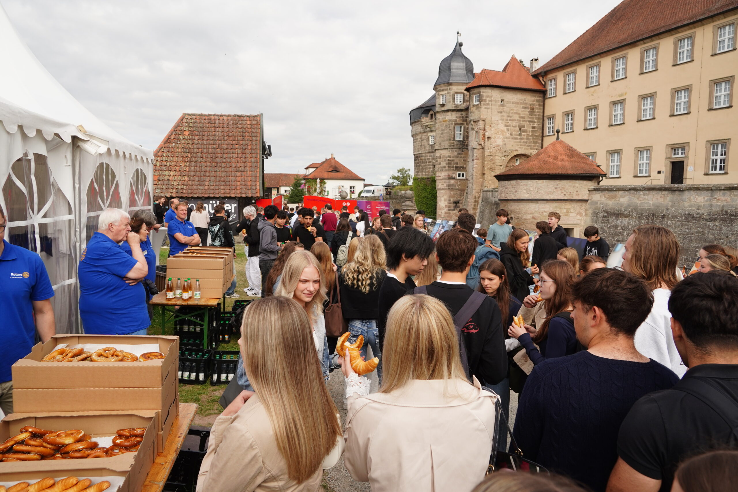 Vor historischen Gebäuden in Kronach versammelt sich eine große Menschenmenge im Freien; die Besucher unterhalten sich und essen Brezeln an Tischen unter einem Zelt. Die Atmosphäre ist lebhaft und festlich, während Jugendliche gemeinsam mit Familien und Freunden das Fest genießen. at Hochschule Coburg