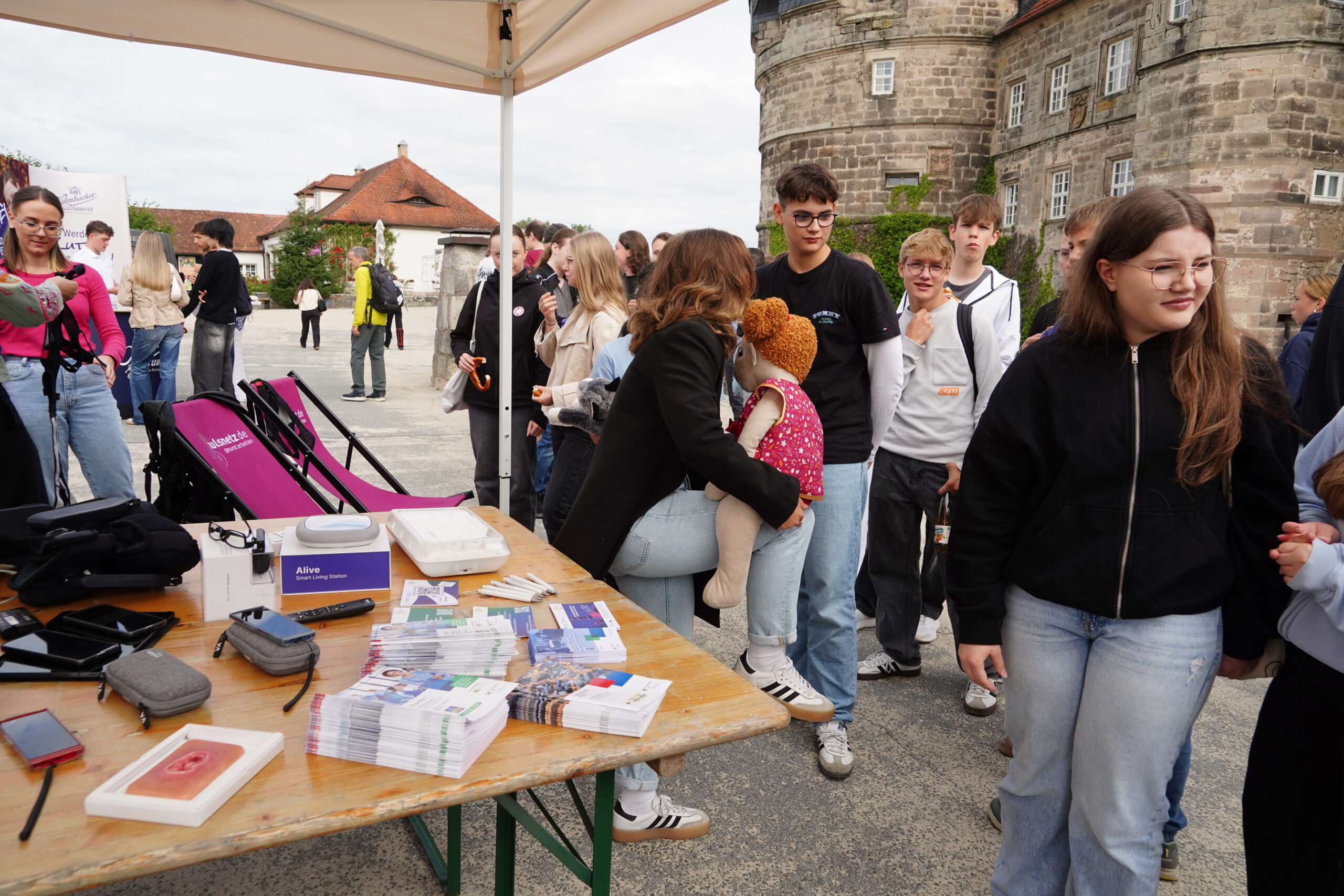 In Kronach versammelt sich eine Gruppe von Menschen, darunter Jugendliche und Erwachsene, im Freien in der Nähe eines Tisches mit Broschüren und medizinischen Hilfsgütern. Eine Person hält eine Puppe in der Hand, während andere vor einem historischen Steingebäude Schlange stehen – eine lebhafte Szene, die an Veranstaltungen mit Persönlichkeiten wie Joey Kelly erinnert. at Hochschule Coburg