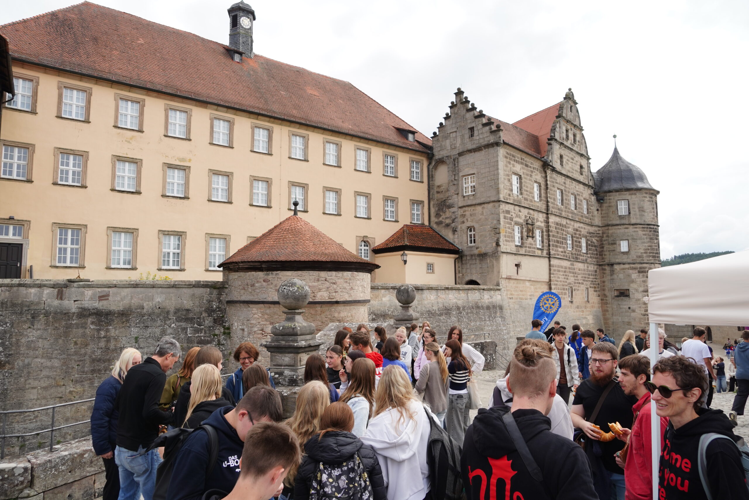 Vor dem historischen Schloss in Kronach mit seinen hellbraunen Mauern und roten Dächern hat sich eine große Menschenmenge versammelt, darunter viele Jugendliche; einige unterhalten sich, andere essen, und im Hintergrund ist ein blaues Rotary-Schild zu sehen. at Hochschule Coburg